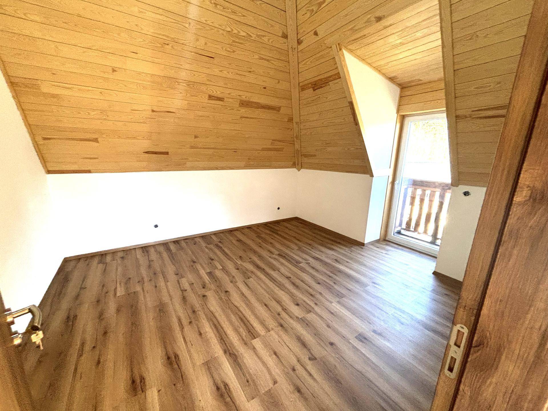 Schlafzimmer mit Holzdachschrägen, Laminatboden und Zugang zu einem kleinen Balkon.