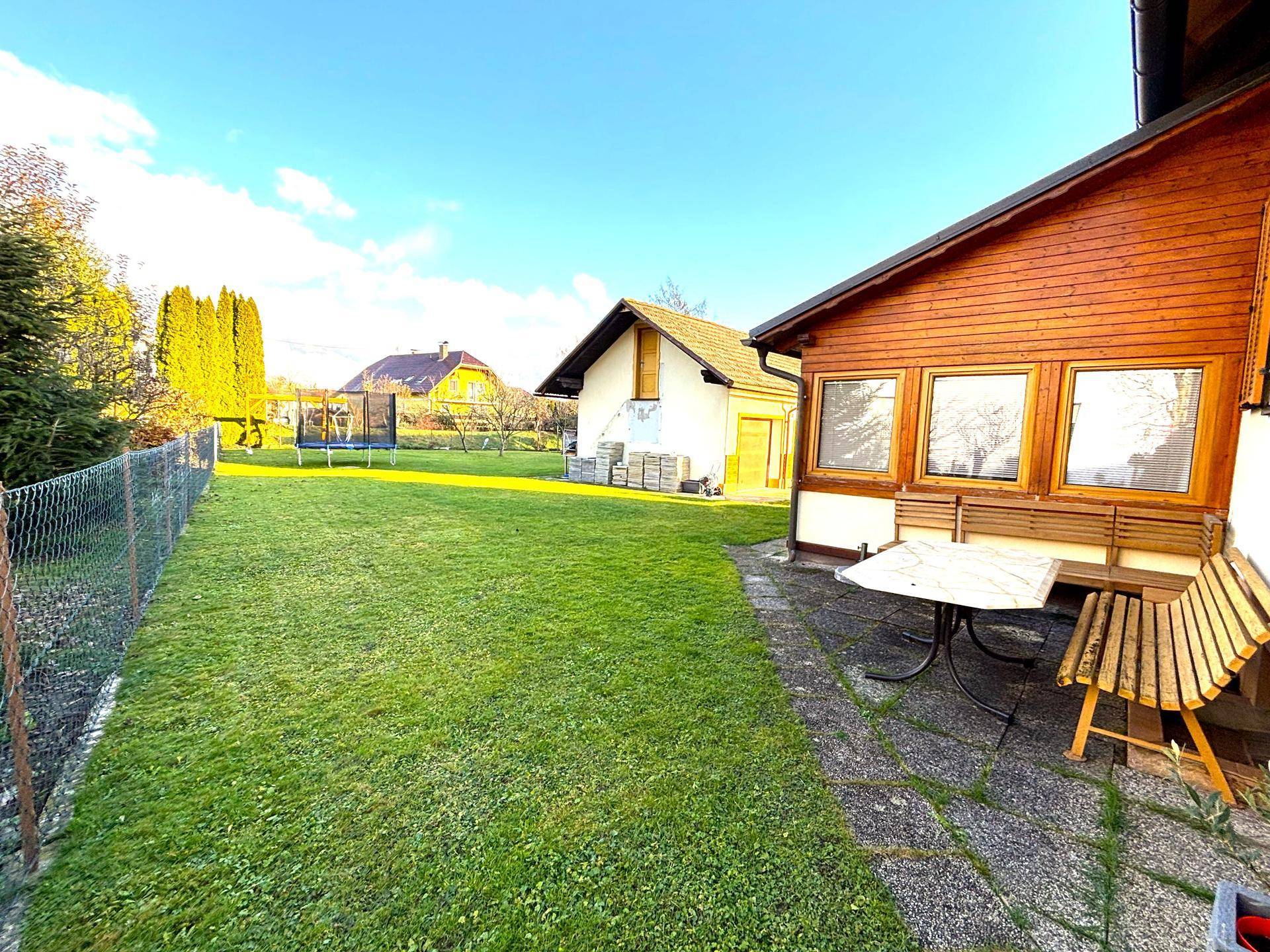 Großer, sonniger Garten mit Rasenfläche, Trampolin und einer kleinen Terrasse mit Sitzgelegenheiten.
