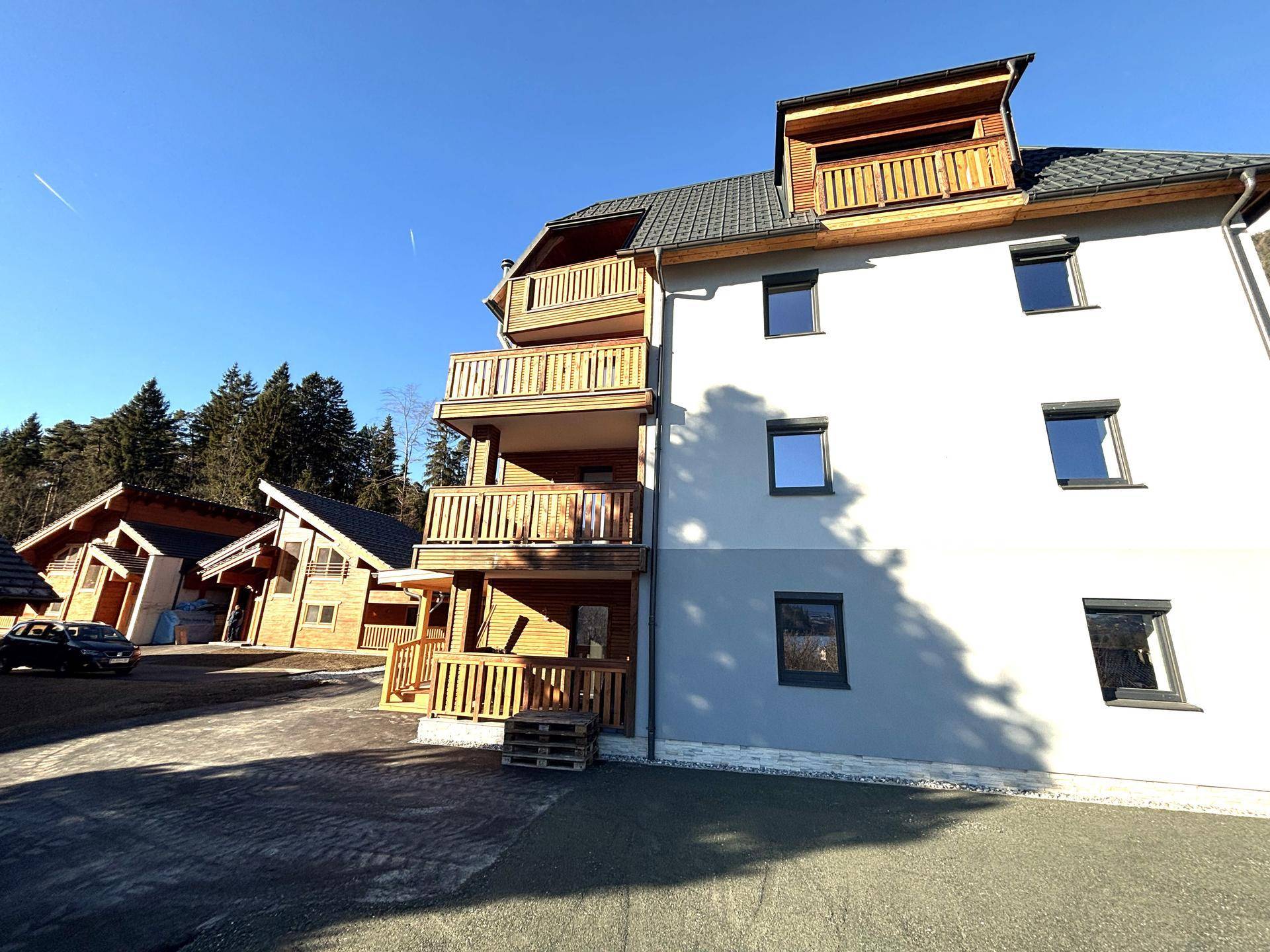 Modernes Gebäude mit hellgrauer Fassade, Holzverkleidung und mehreren Balkonen in ländlicher Umgebung.