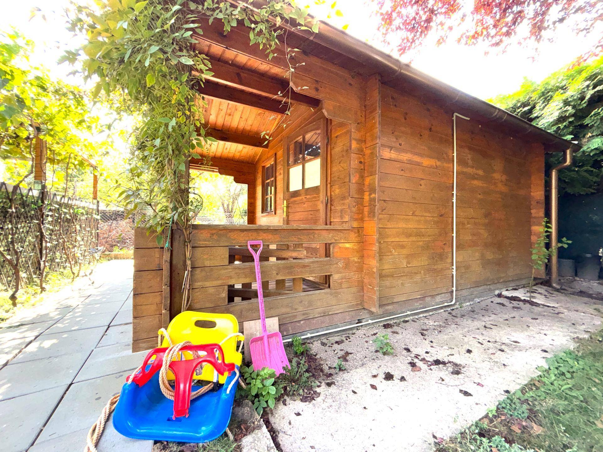Kleines Holzhaus im Garten mit Veranda und Spielzeug davor, umgeben von Bäumen.
