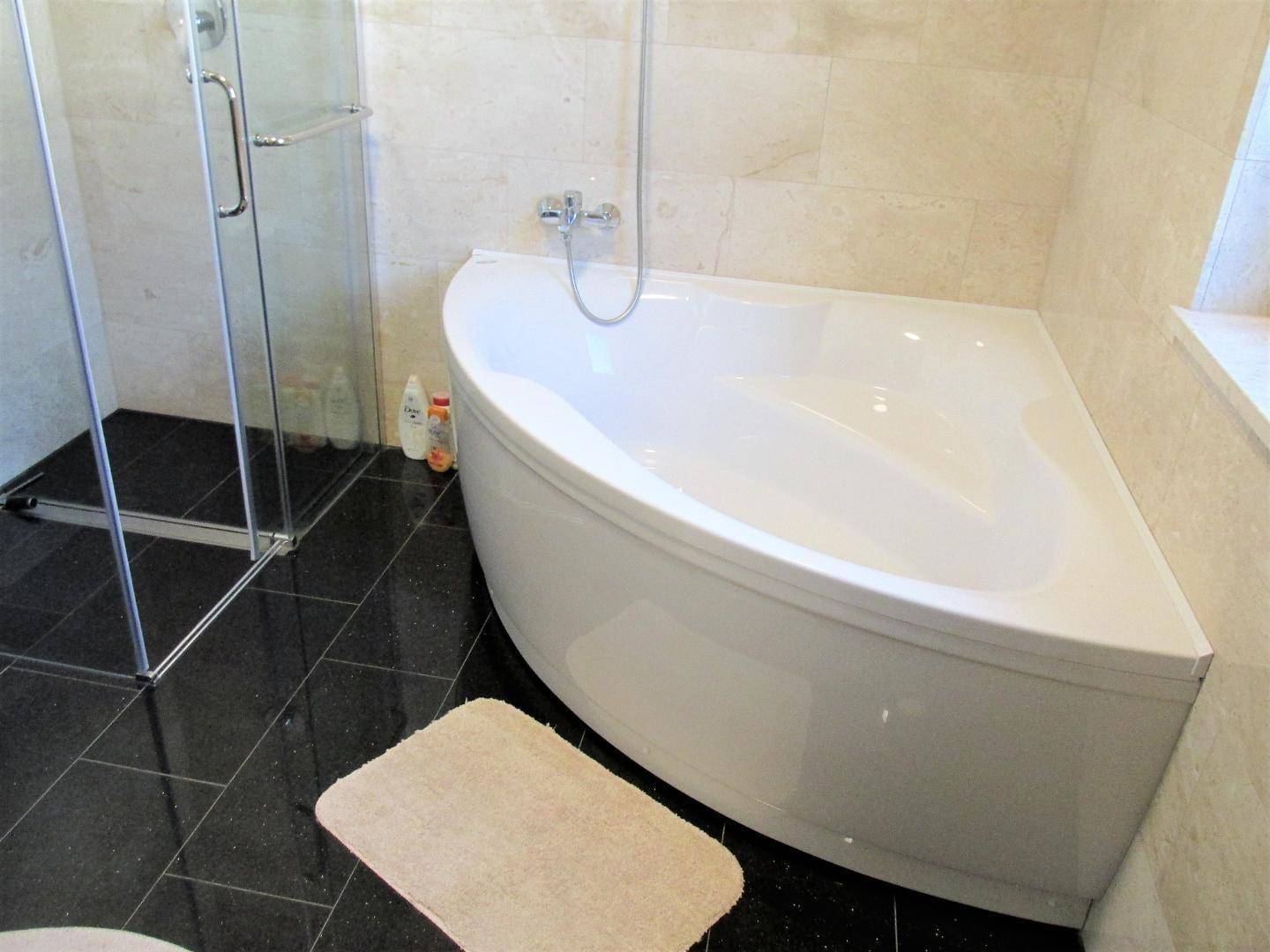 Geräumiges Badezimmer mit Eckbadewanne und separater Glasdusche auf dunklem Fliesenboden.