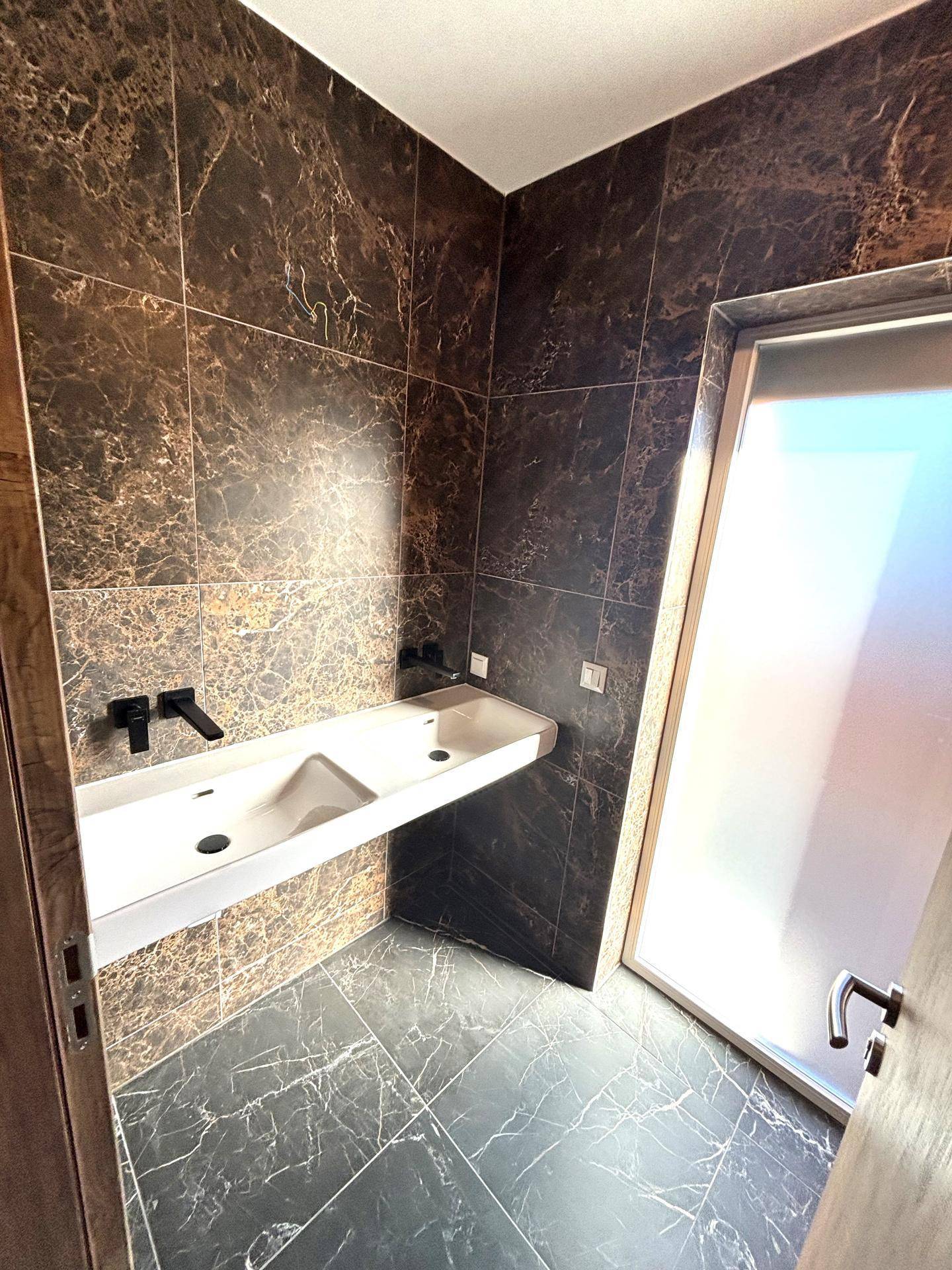 Modernes Badezimmer mit dunklen Marmorfliesen, Doppelwaschbecken und Duschzugang.