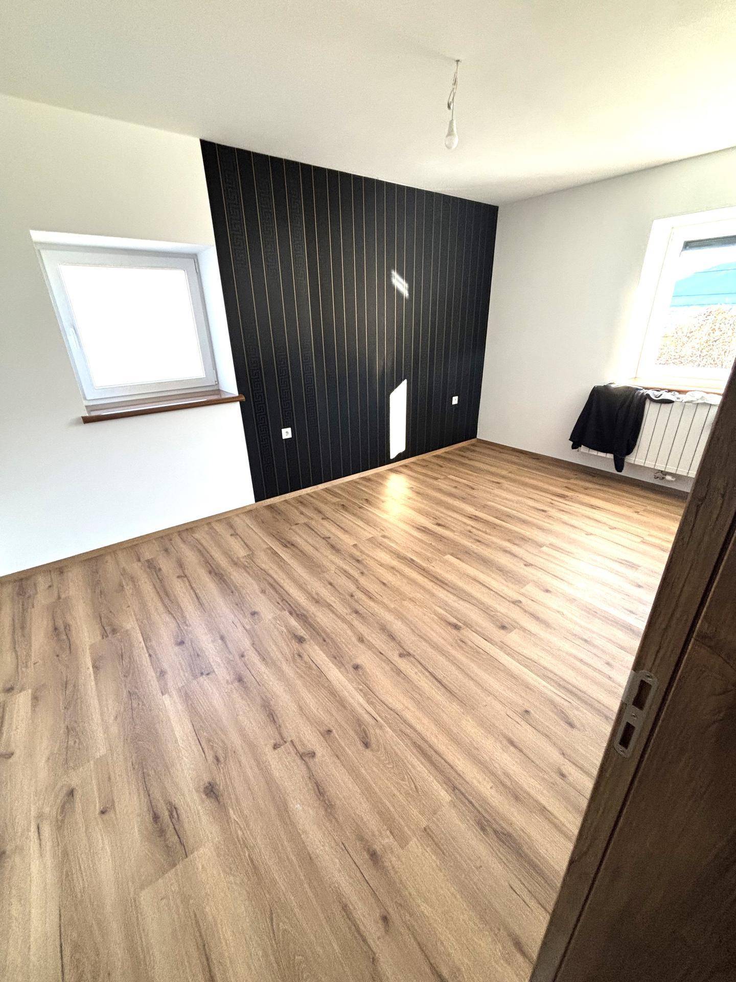 Schlafzimmer mit Laminatboden, einer schwarzen Akzentwand und zwei Fenstern für viel Tageslicht.