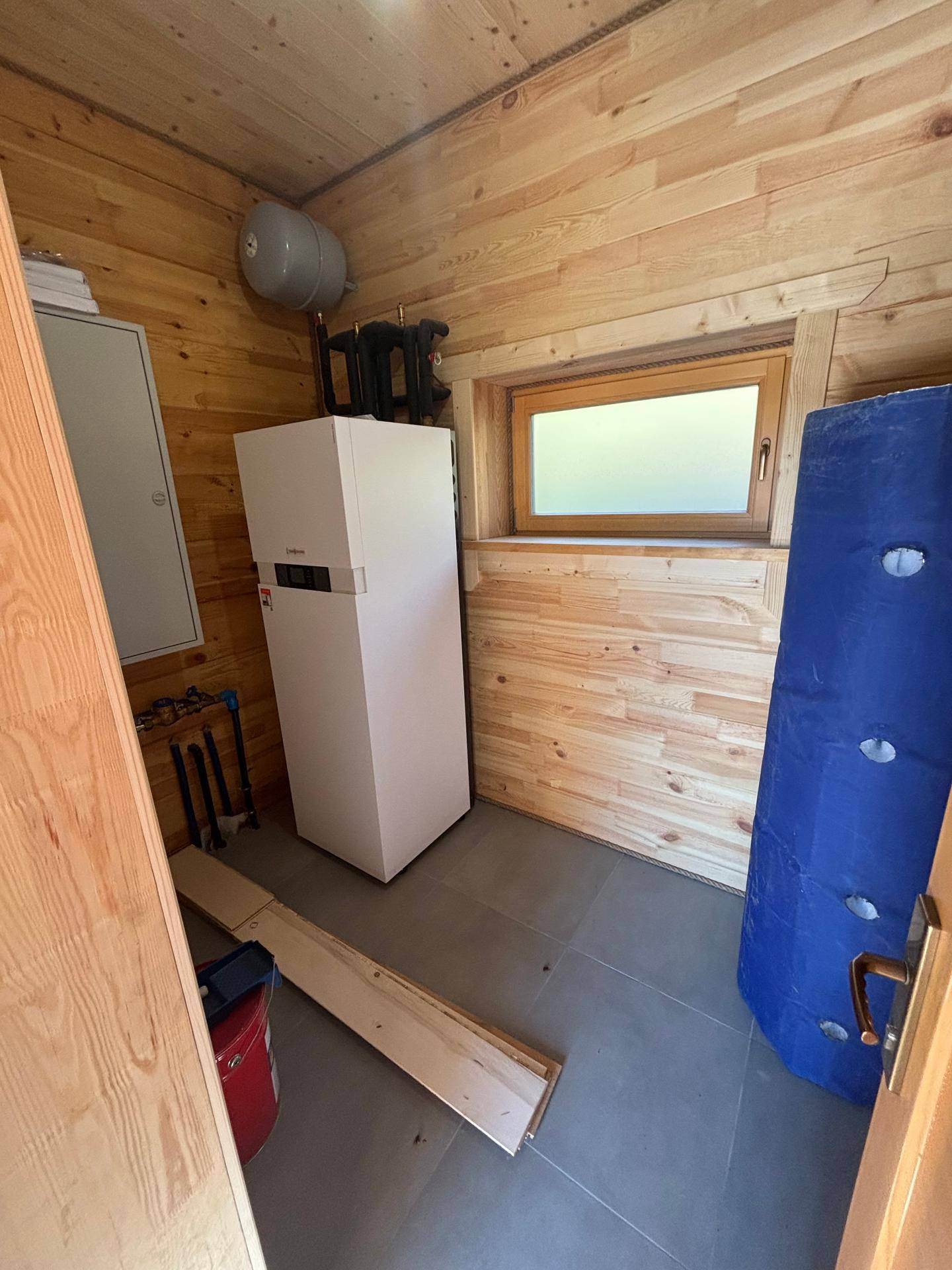 Hauswirtschaftsraum mit Wärmepumpe, Fenster und Fliesenboden in einem Holzhaus.