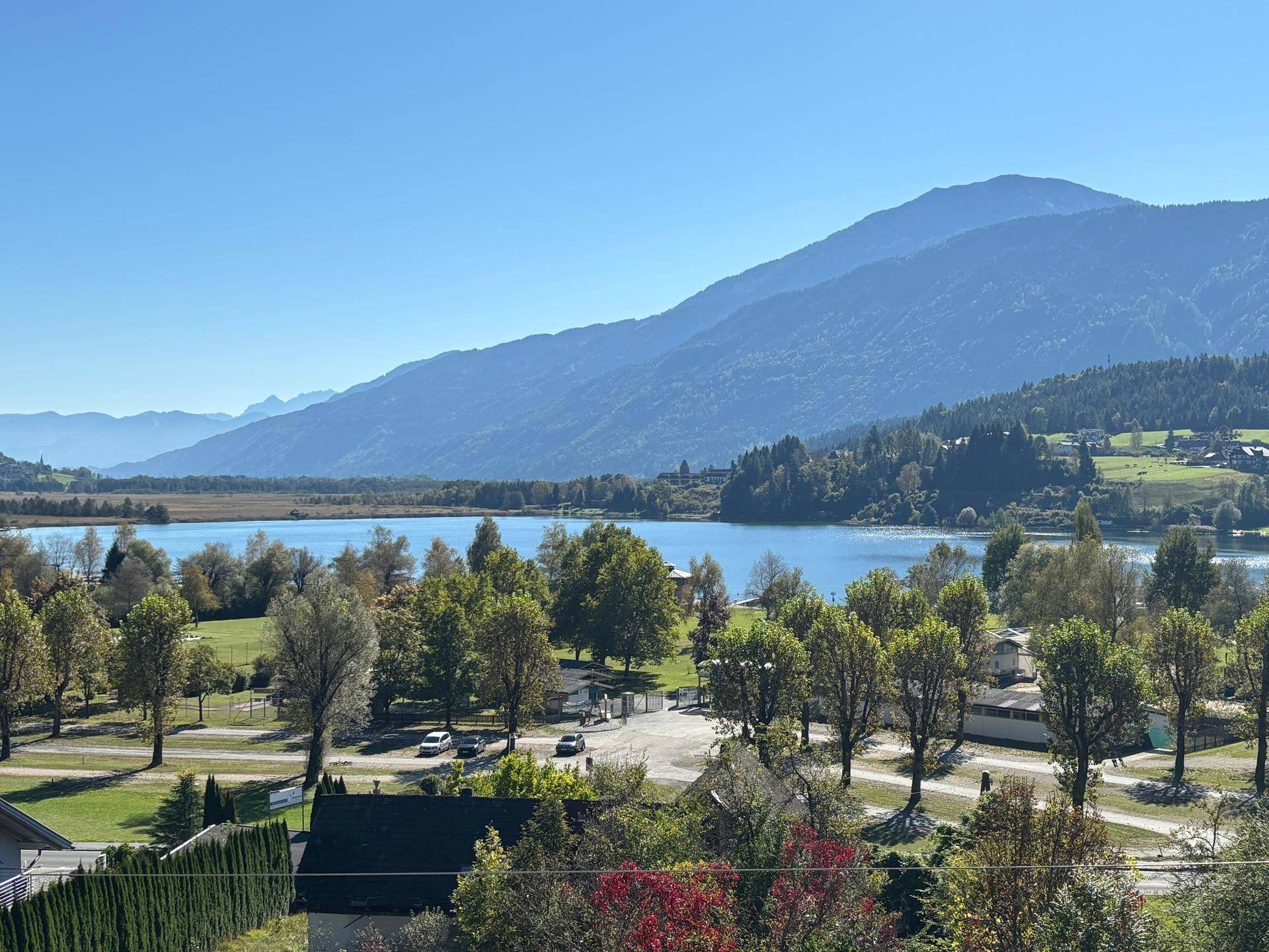 Malerischer Ausblick auf einen ruhigen See, umgeben von grünen Hügeln und majestätischen Bergen unter blauem Himmel.