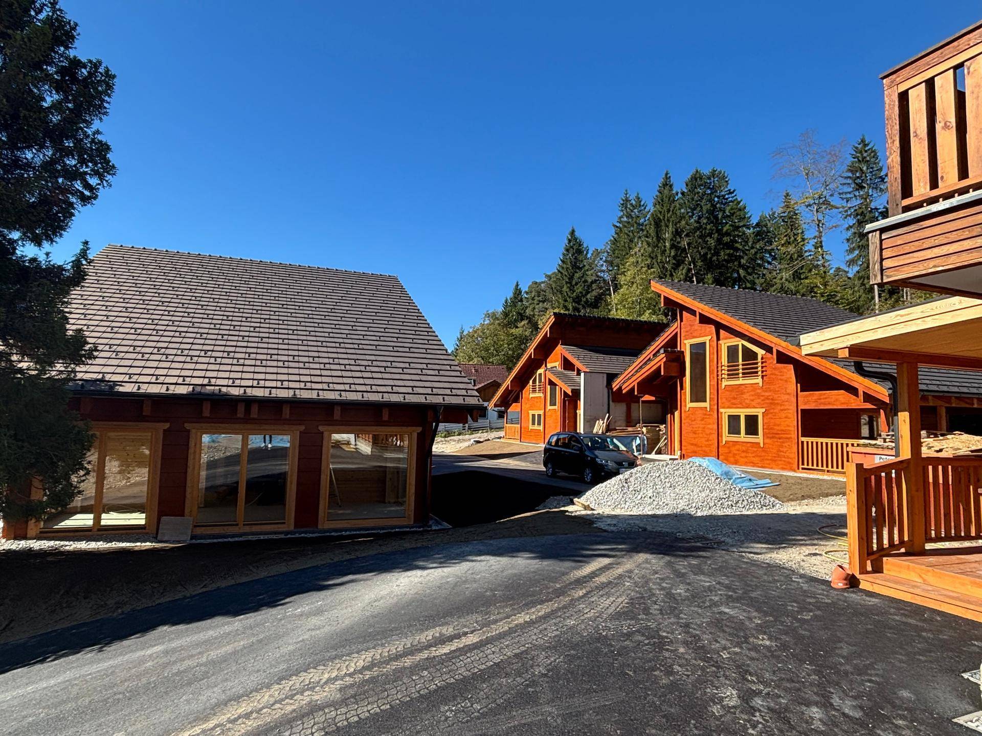 Drei neue Holzhäuser im Bau, umgeben von Wald und Bergen, mit asphaltierter Zufahrt.