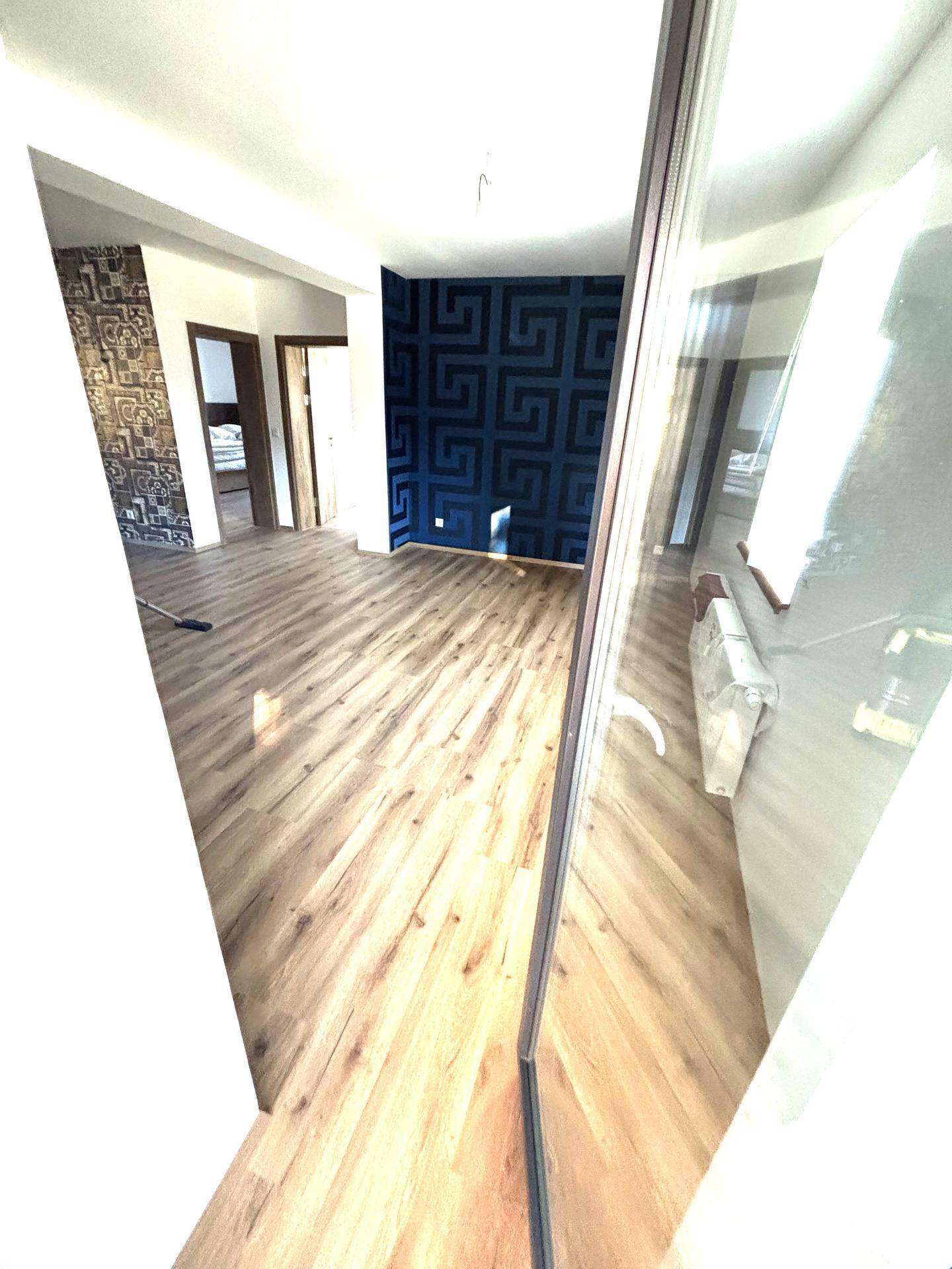 Helles Wohnzimmer mit Laminatboden und einer blauen geometrischen Akzentwand, durch Glastür sichtbar.