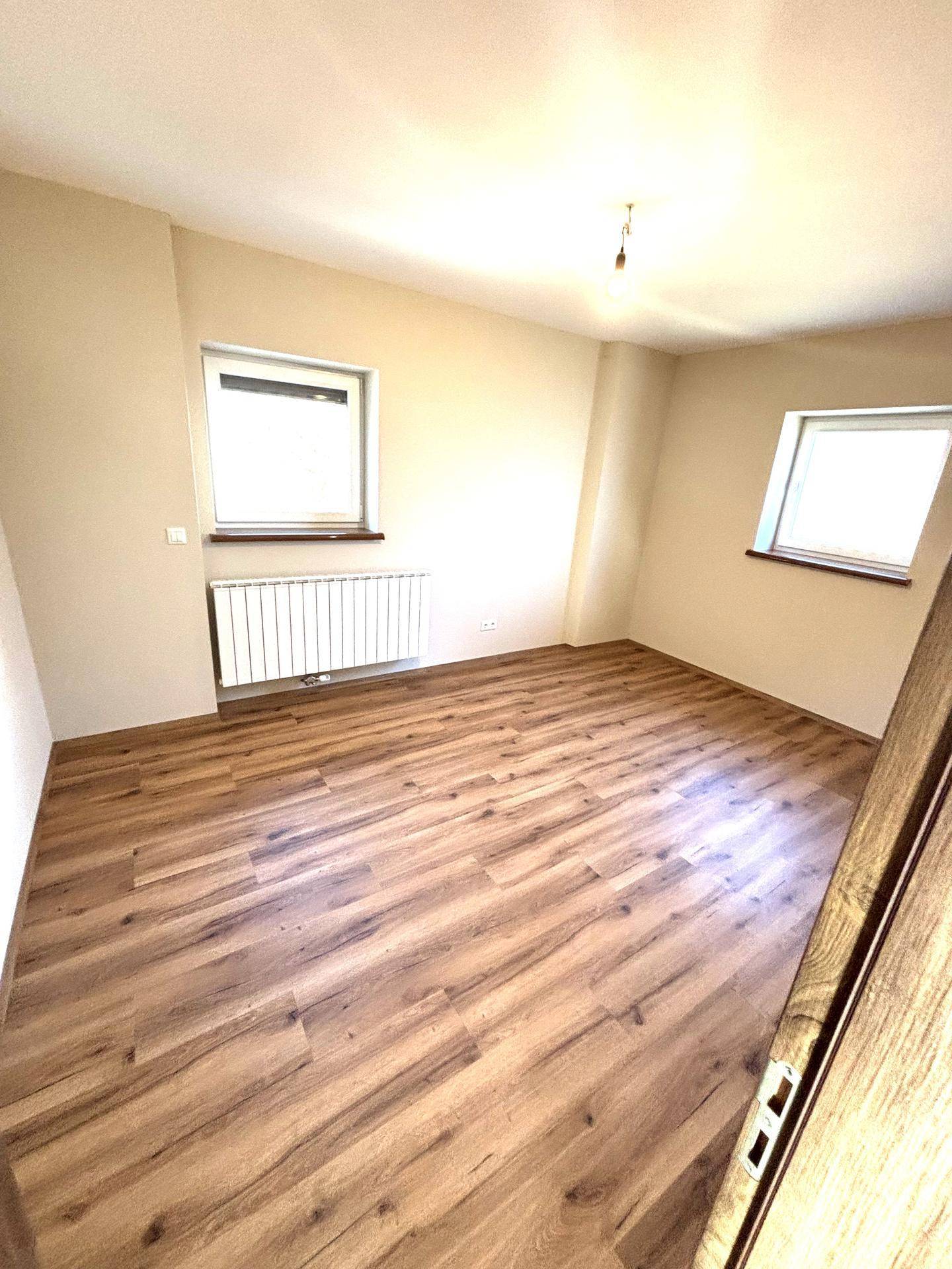 Helles Schlafzimmer mit Laminatboden, zwei Fenstern und einem Heizkörper.