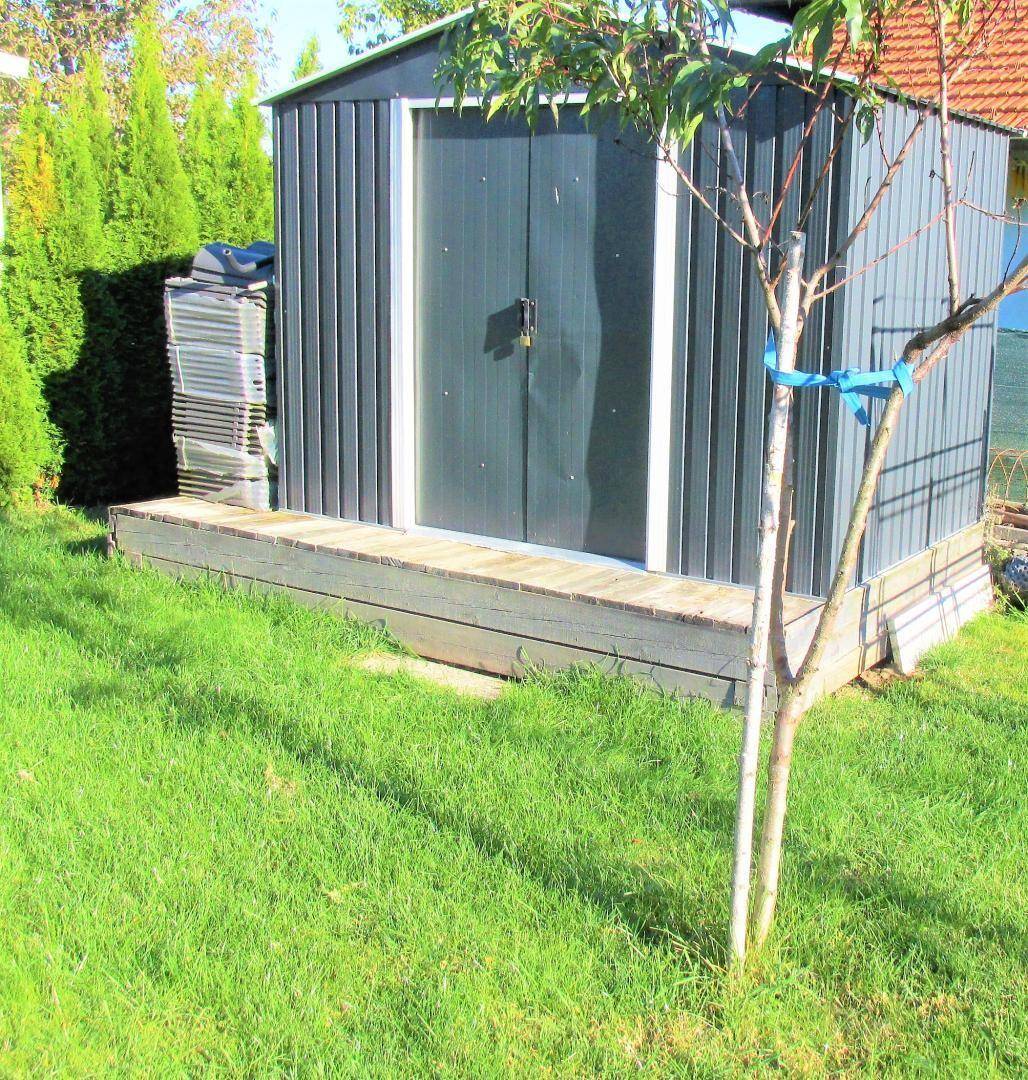 Metall-Gartenschuppen mit kleiner Holzterrasse und umgebender Rasenfläche.