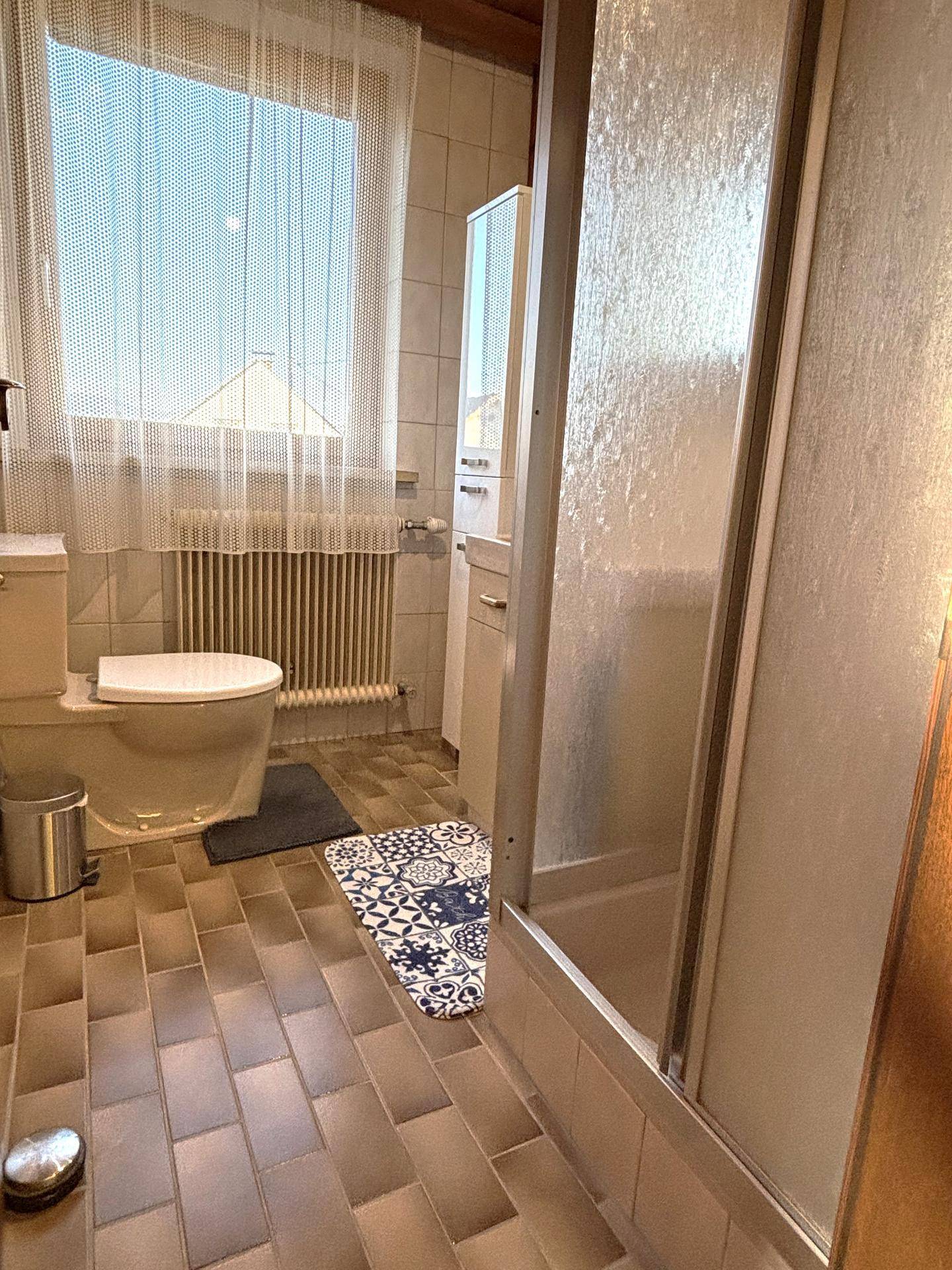 Badezimmer mit Duschkabine, WC und Fenster, ausgestattet mit beigefarbenen Fliesen.