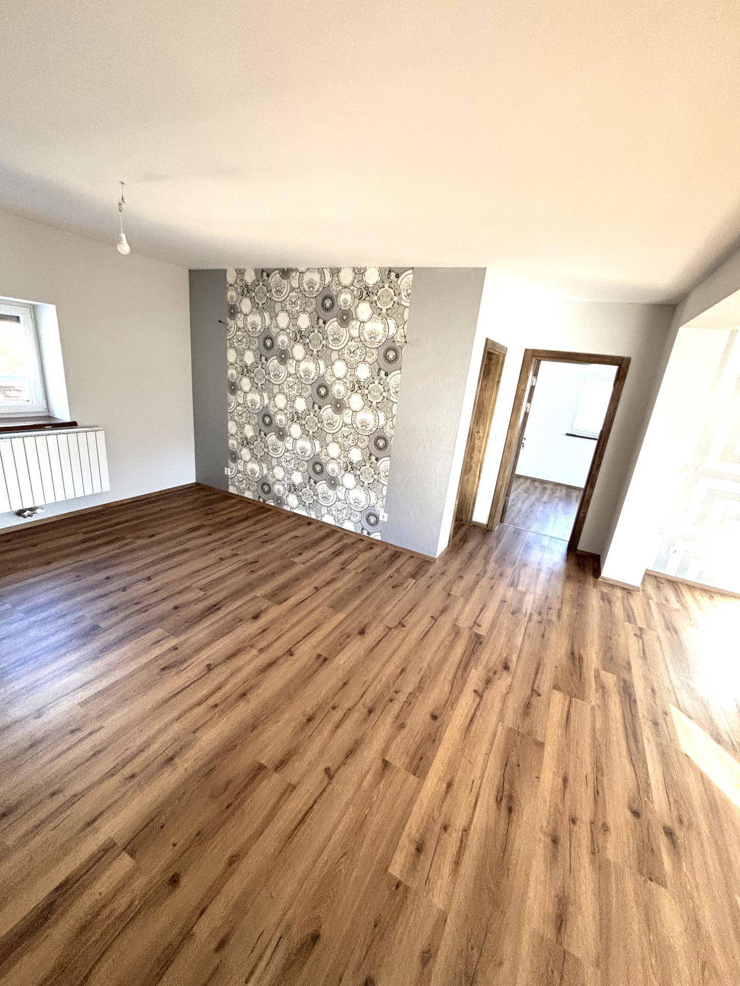 Geräumiges Wohnzimmer mit Laminatboden und einer auffälligen Mustertapete an der Akzentwand.
