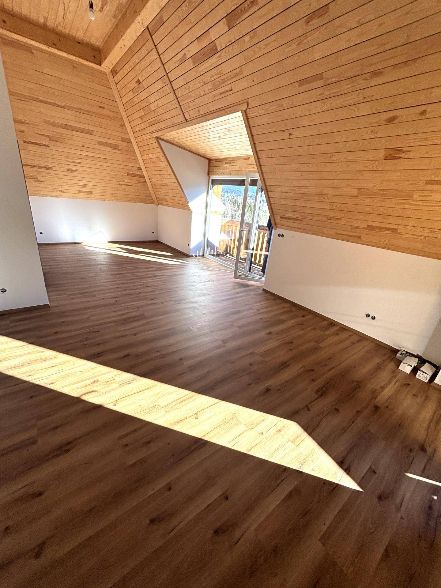 Heller Wohnbereich mit Holzdachschrägen, Laminatboden und Zugang zu einem Balkon.
