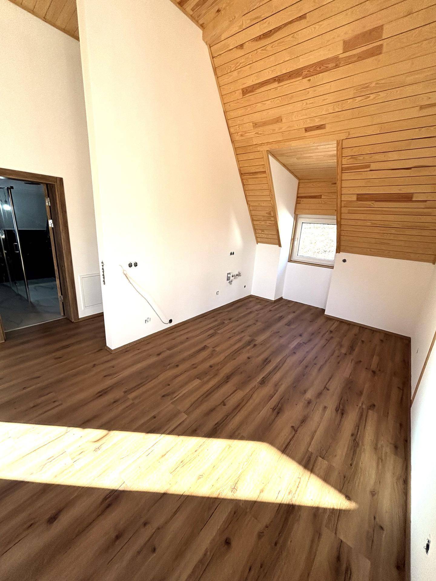Heller Wohnbereich mit Holzdachschrägen, Laminatboden und einem Fenster mit Ausblick.