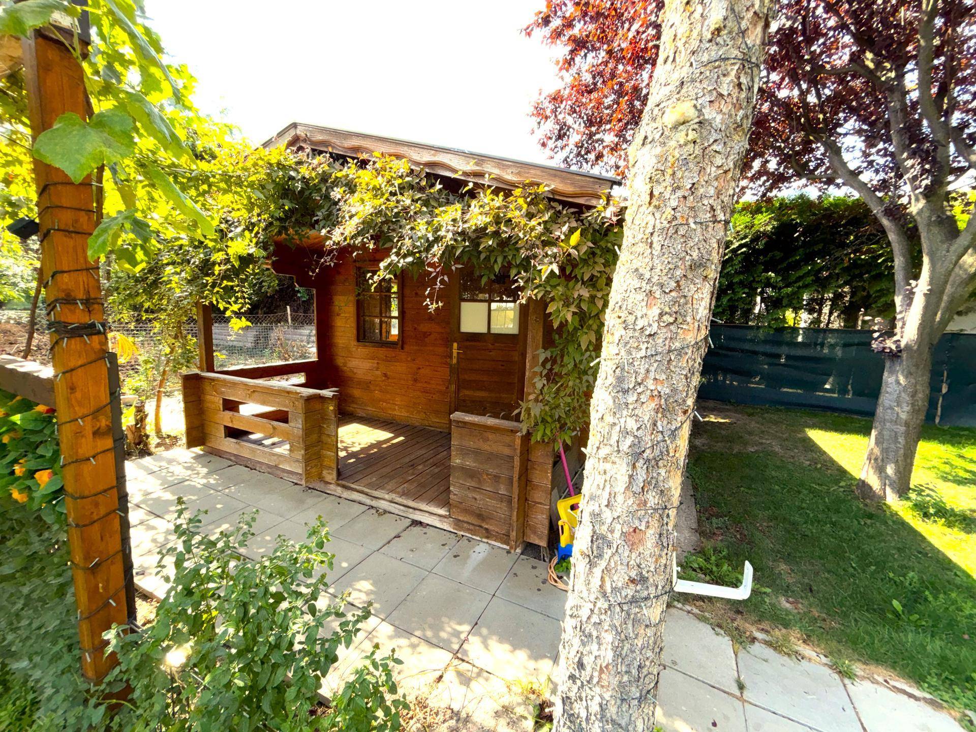 Holzhaus mit Veranda im Garten, umgeben von Bäumen und Sträuchern.