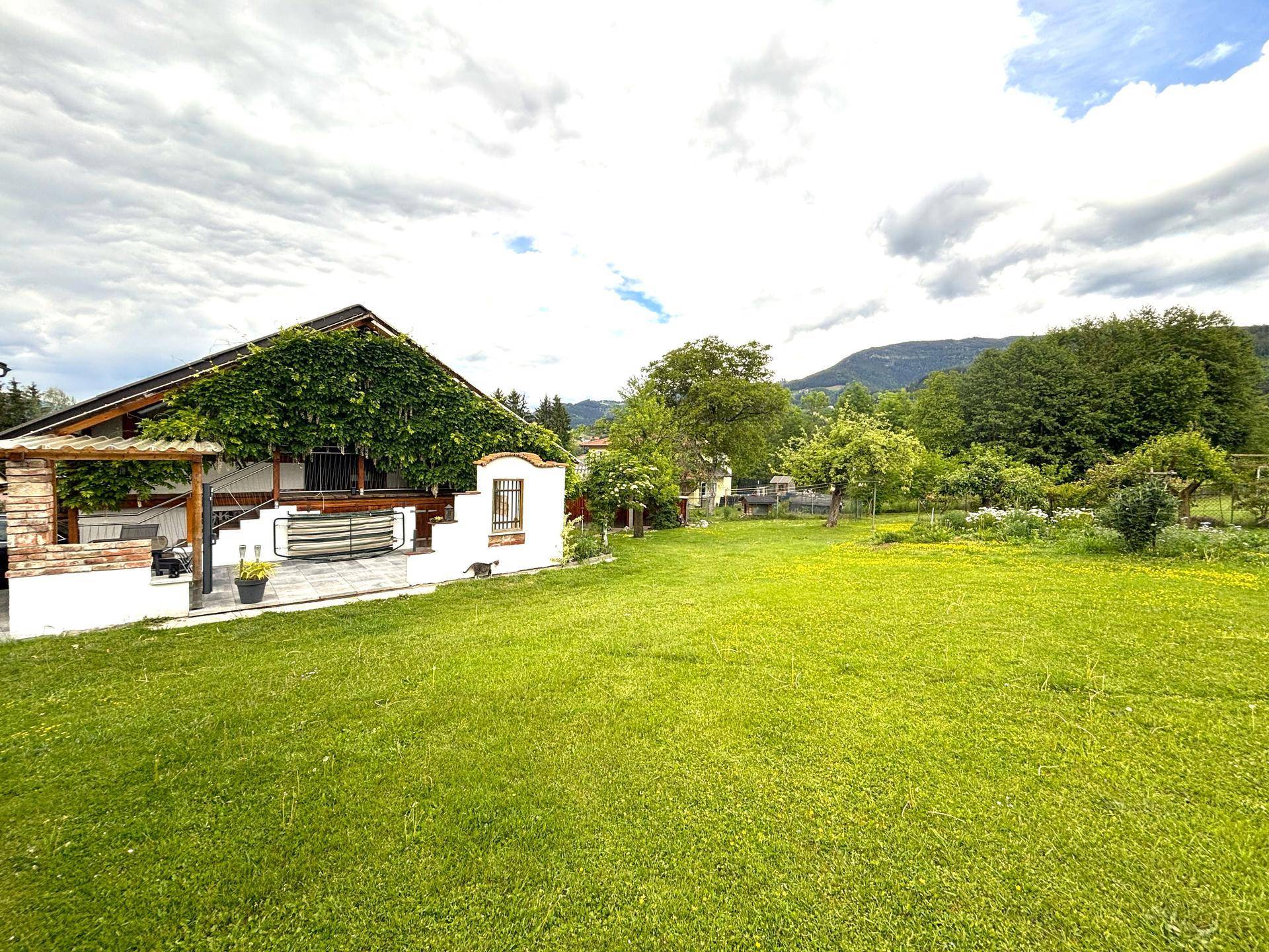 Weitläufiger grüner Garten mit Rasenfläche, Bäumen und einem Haus mit bewachsener Fassade im Hintergrund.