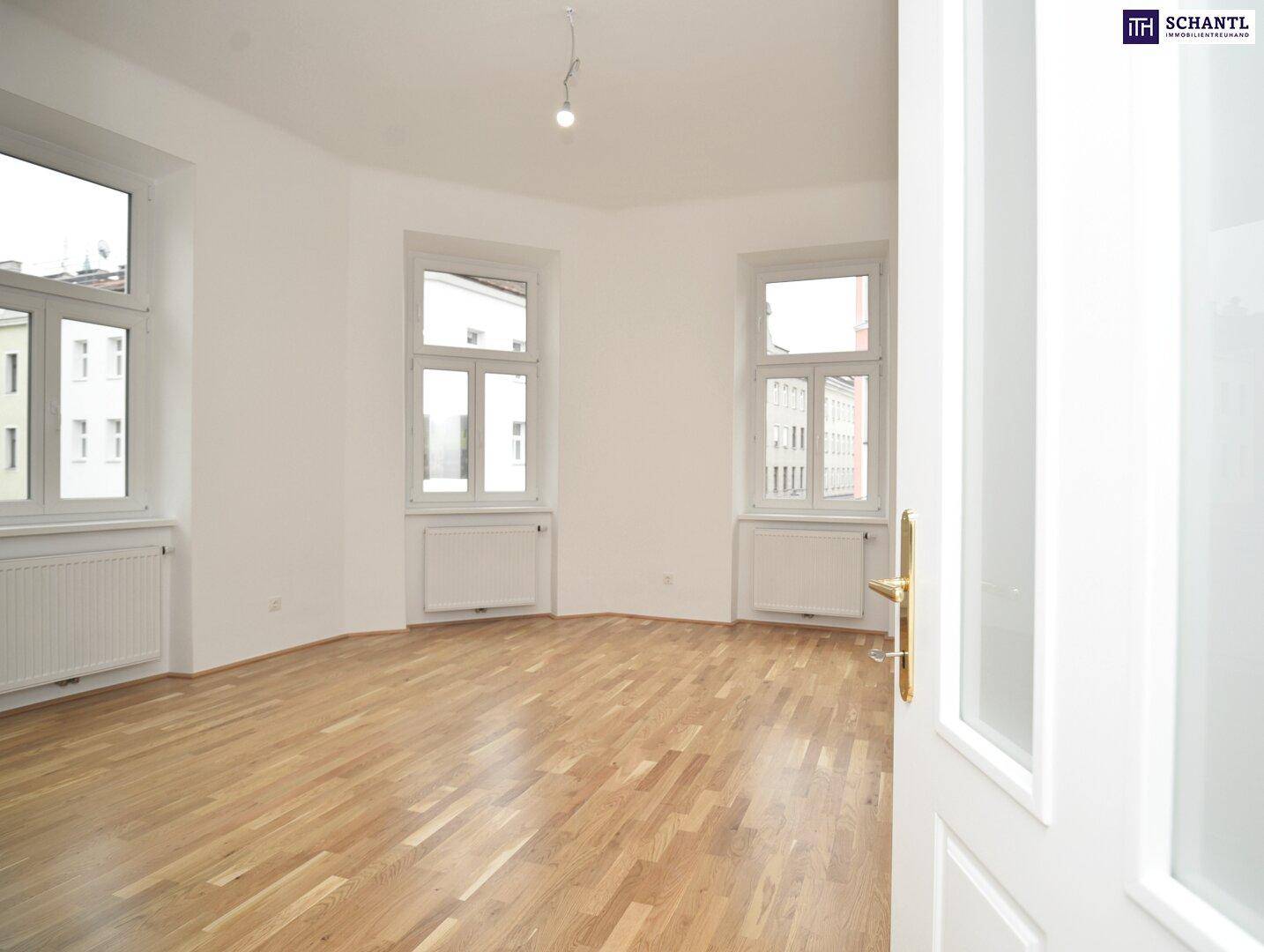 Geräumiger, heller Raum mit Holzboden und großen Fenstern, ideal als Wohnbereich.