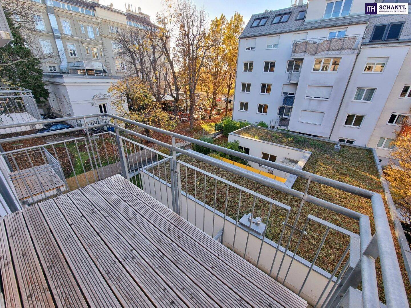 Balkon mit Holzdeck und Blick auf ein begrüntes Dach sowie umliegende Gebäude.