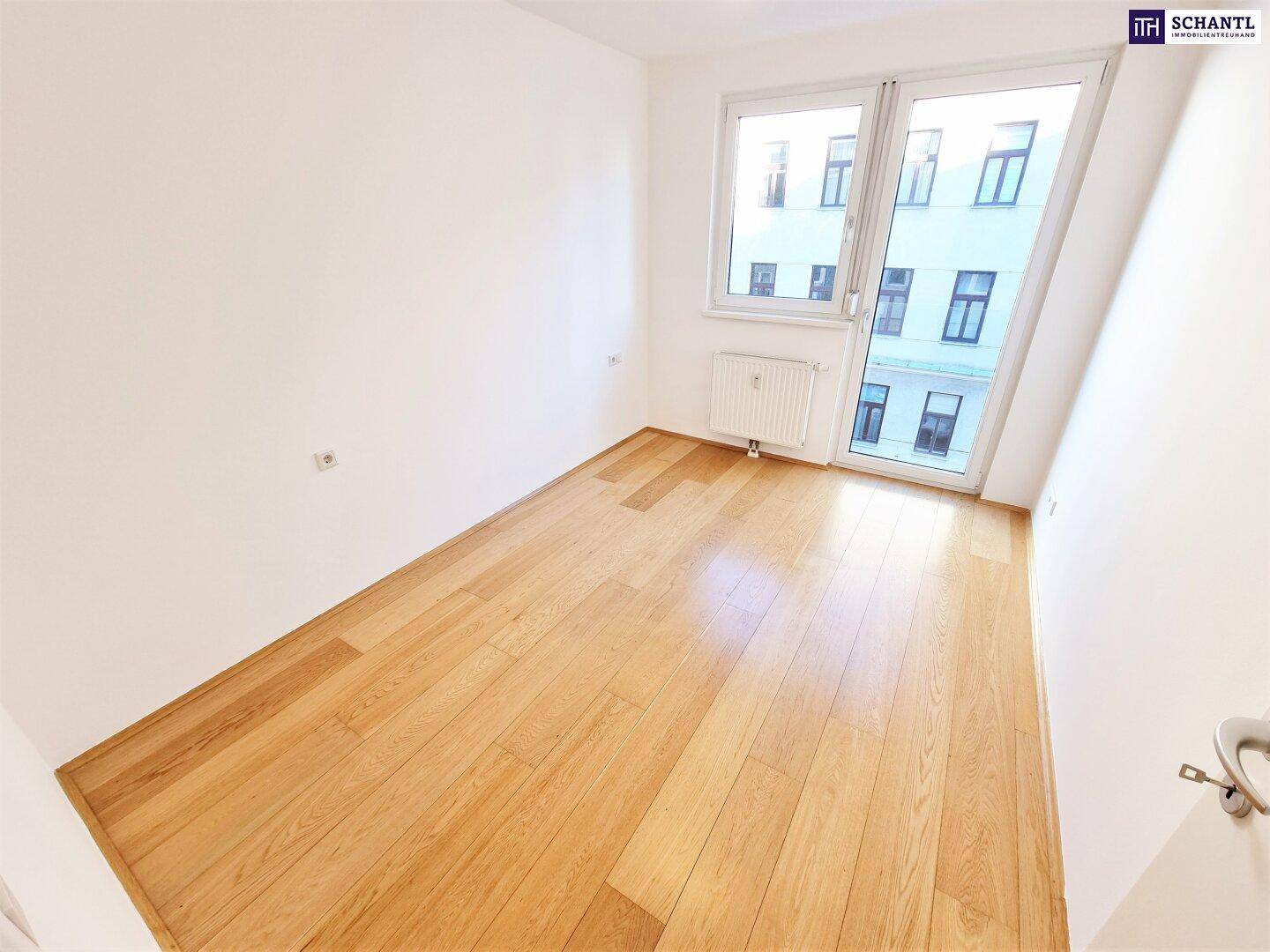 Helles Zimmer mit Holzboden und Fensterfront, die Zugang zu einem Balkon ermöglicht.