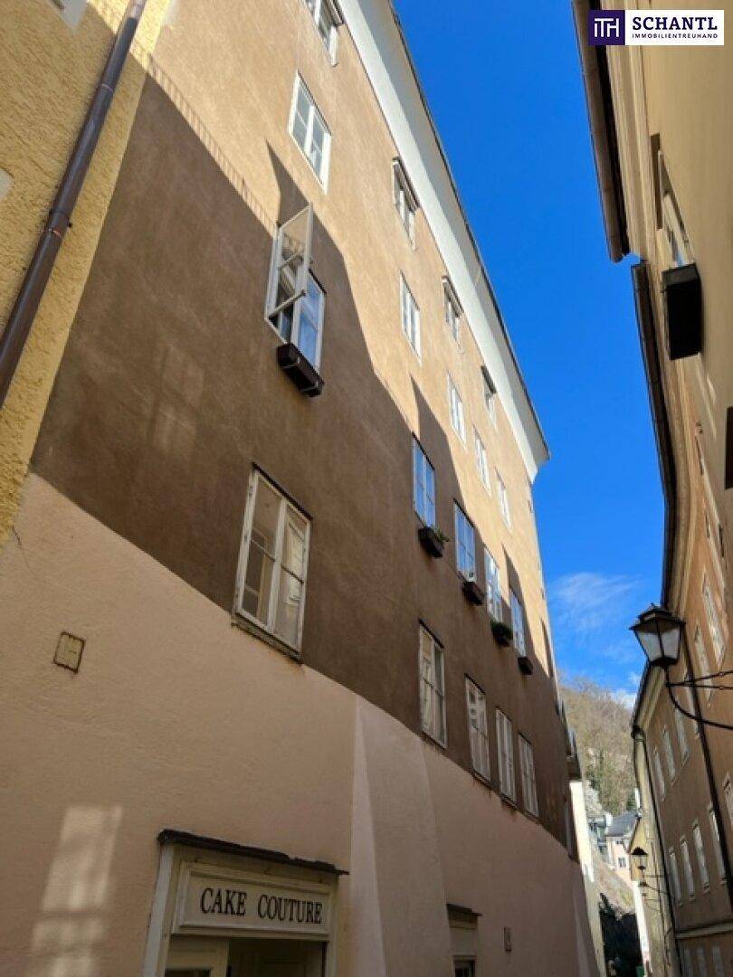 Historische Fassade eines Gebäudes in einer engen Gasse mit vielen Fenstern unter blauem Himmel und Bergblick.