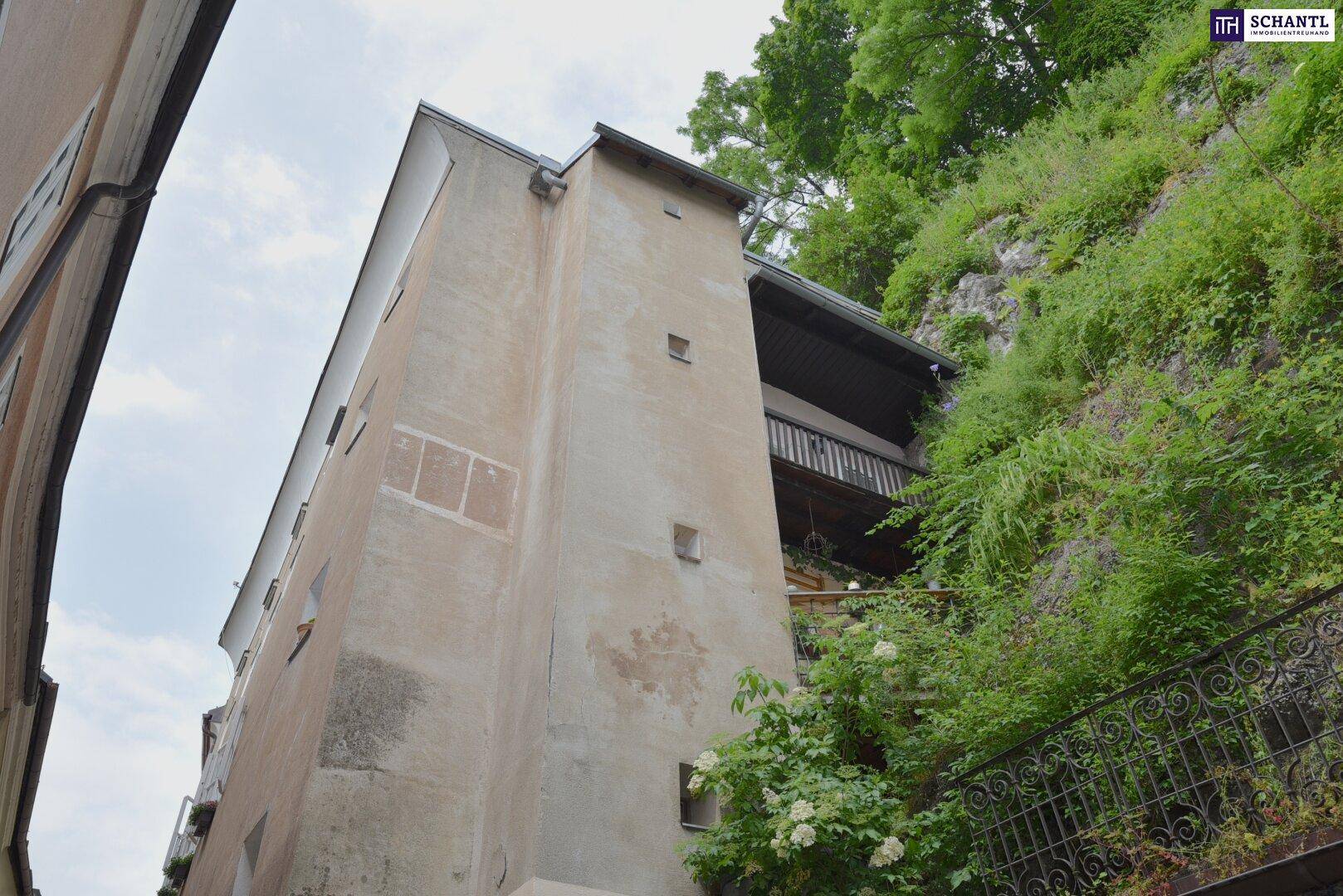 Seitenansicht eines alten Gebäudes mit Balkonen, umgeben von üppiger Vegetation und Felsen.