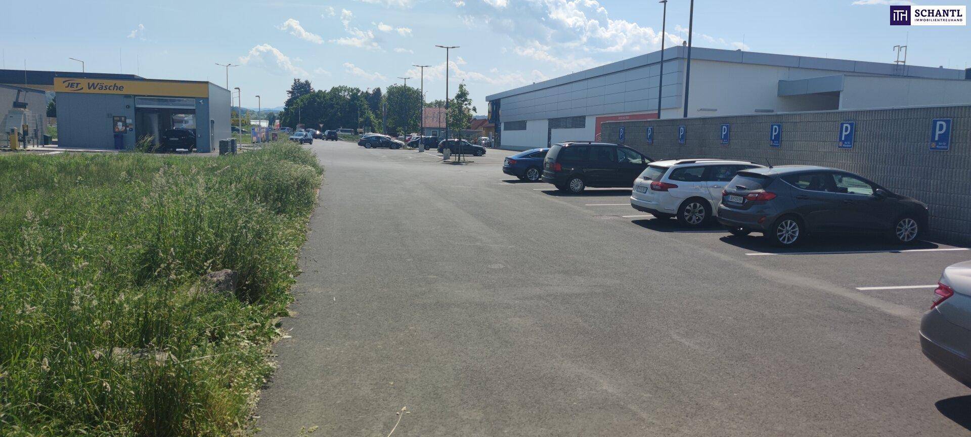 Außenansicht eines großen Parkplatzes neben einem modernen Gewerbegebäude mit einer Autowaschanlage im Hintergrund.