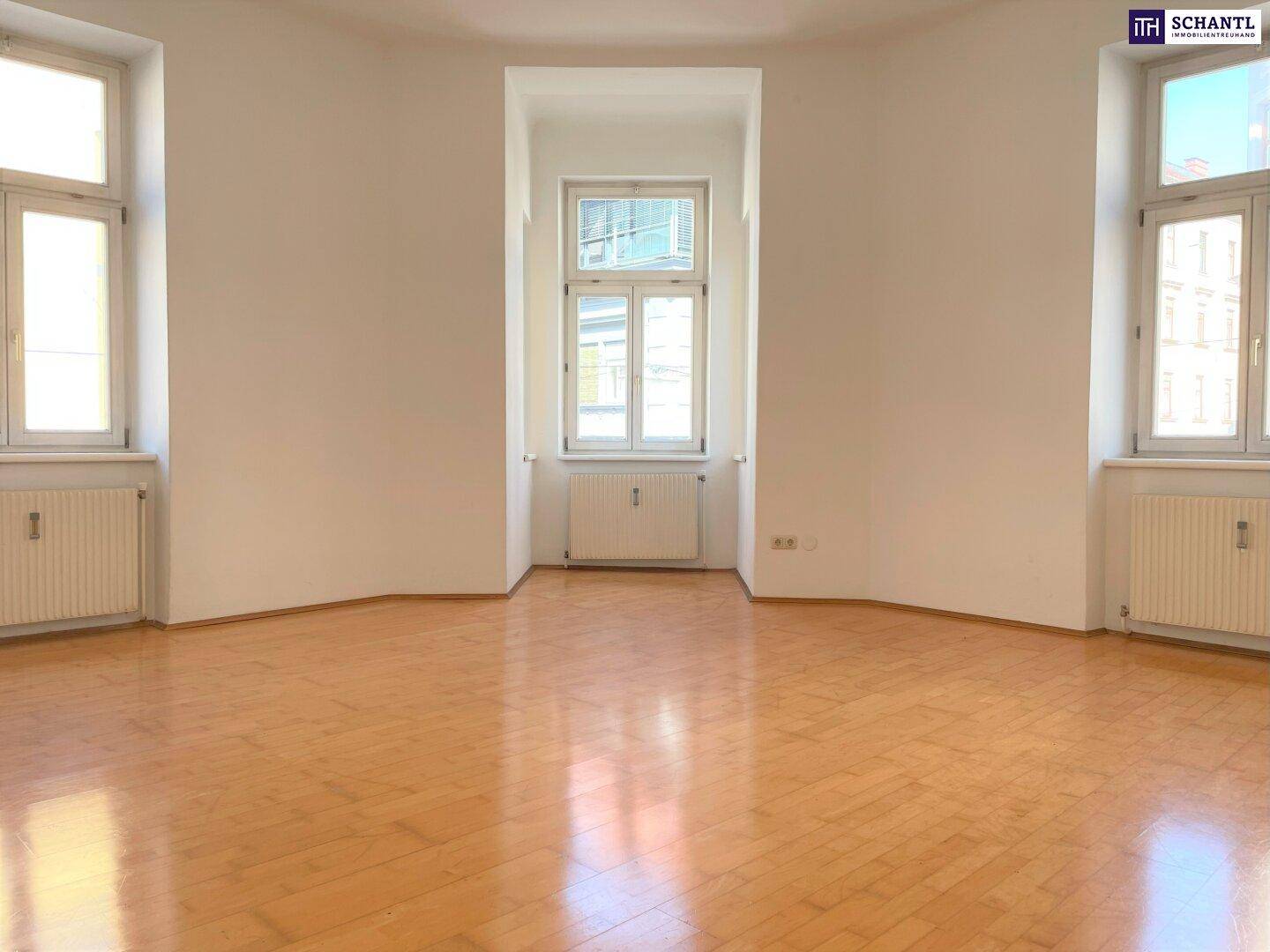 Helles Zimmer mit Fischgrätparkettboden, weißen Wänden und drei Fenstern mit Heizkörpern.