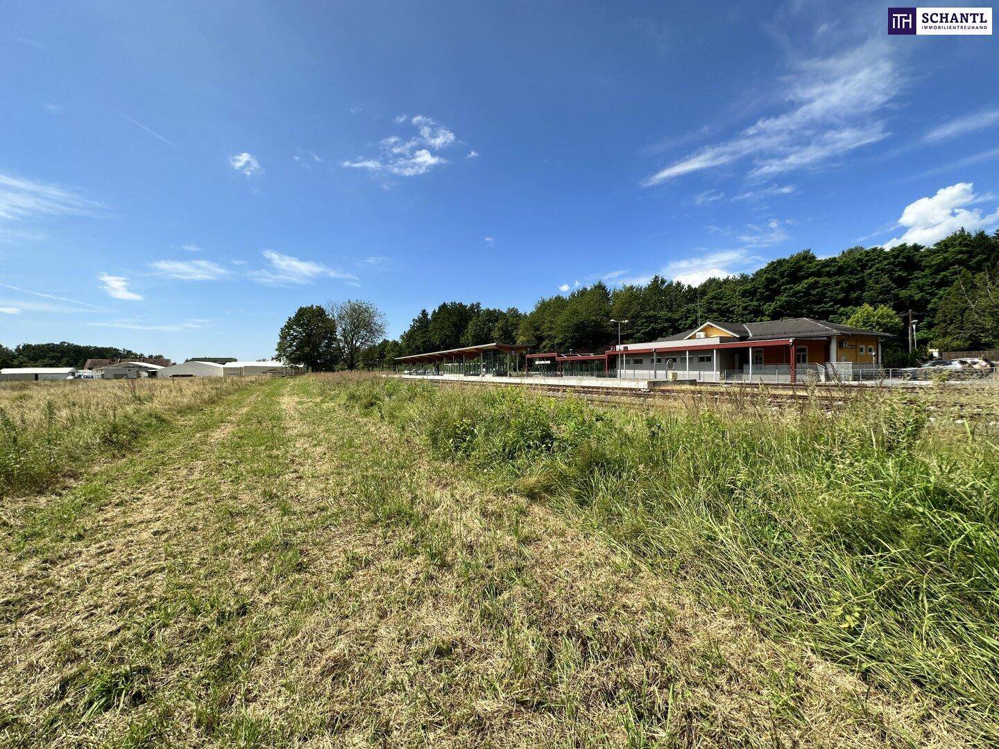 Grasbewachsenes Gelände mit einem Bahnhof und Gleisen im Mittelgrund, umgeben von Bäumen und Hügeln.