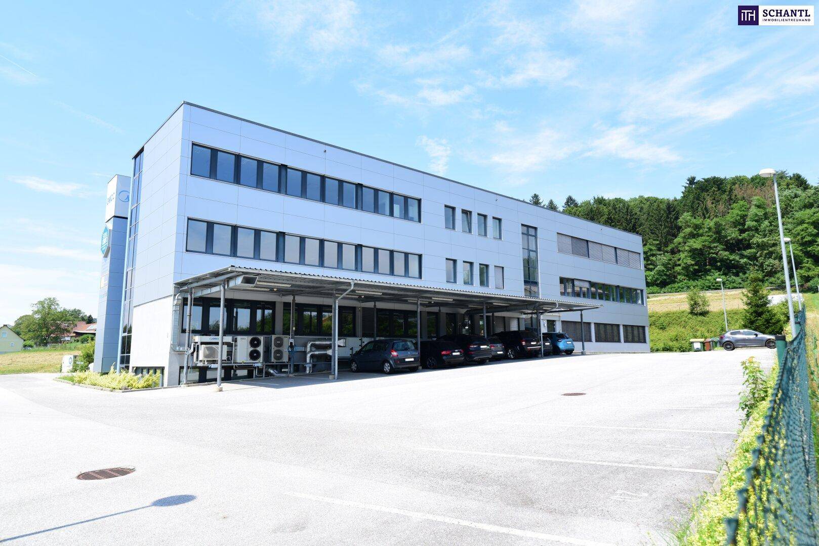 Gesamtansicht des Bürogebäudes mit großem Parkplatz und angrenzender Grünfläche unter blauem Himmel.