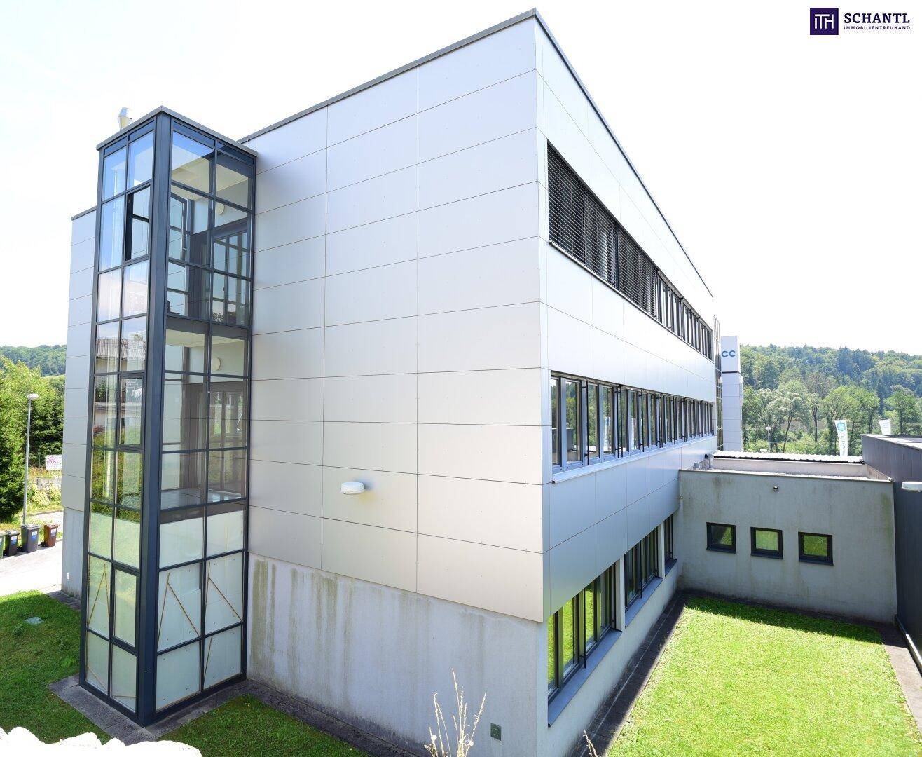 Ansicht des modernen Bürogebäudes mit Glasaufzug und gepflegter Rasenfläche an einem sonnigen Tag.