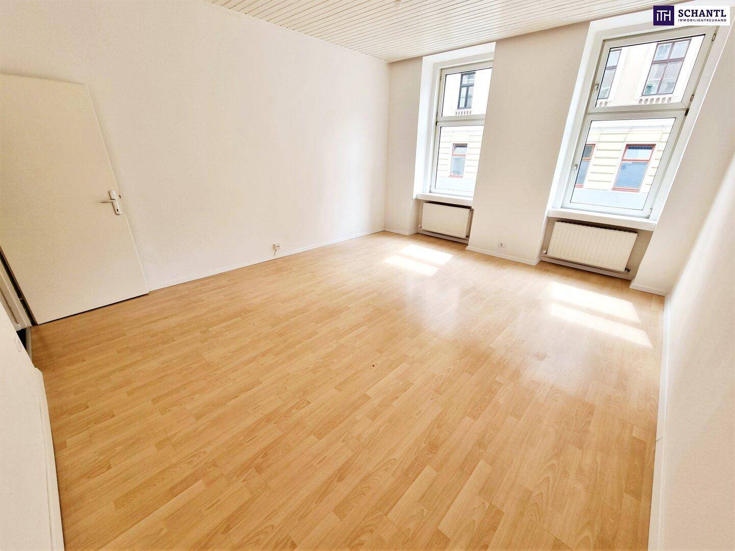 Helles Zimmer mit Holzfußboden und großen Fenstern, die viel Tageslicht hereinlassen.