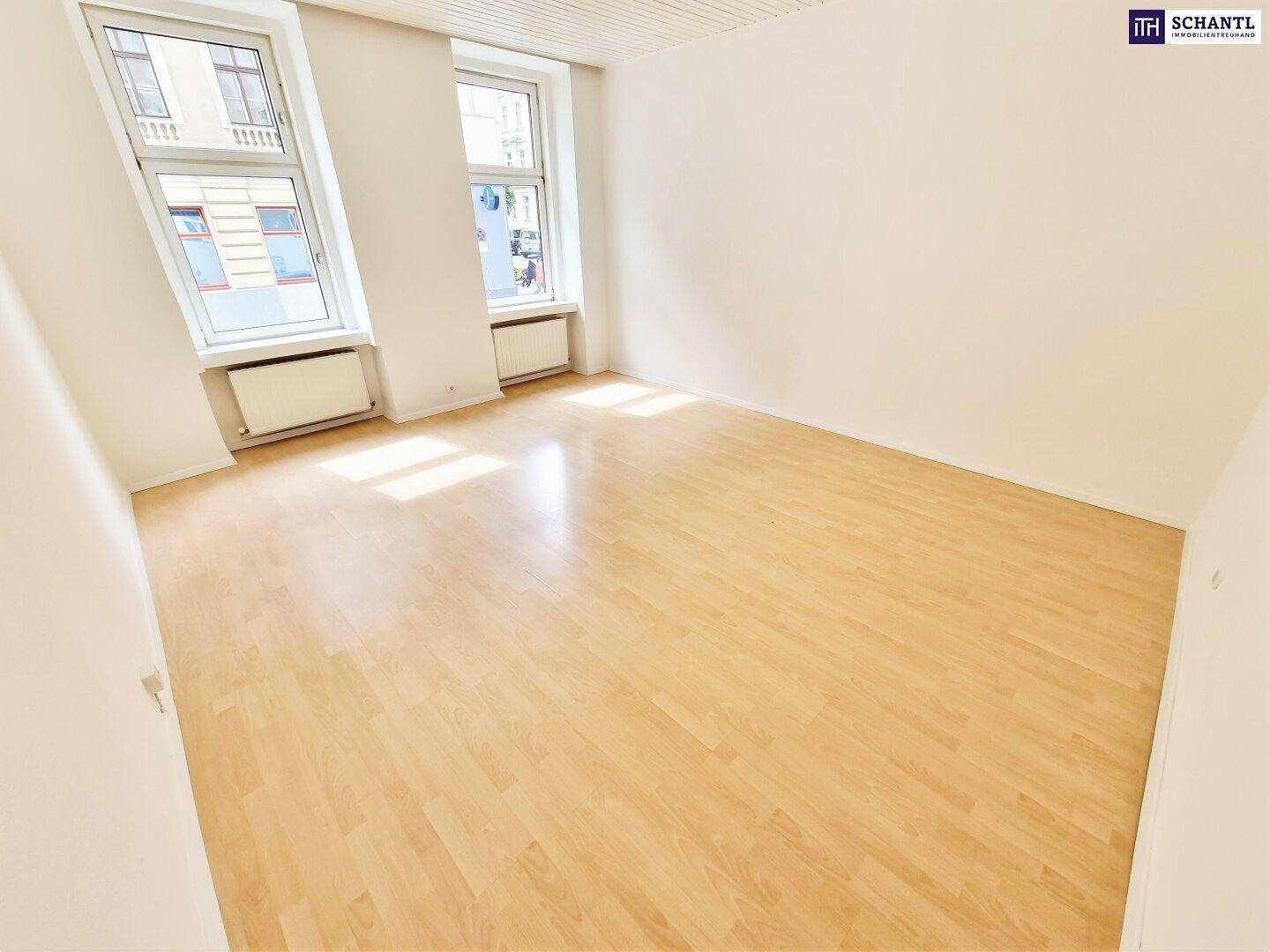 Helles Zimmer mit Holzfußboden und großen Fenstern, die viel Tageslicht hereinlassen.
