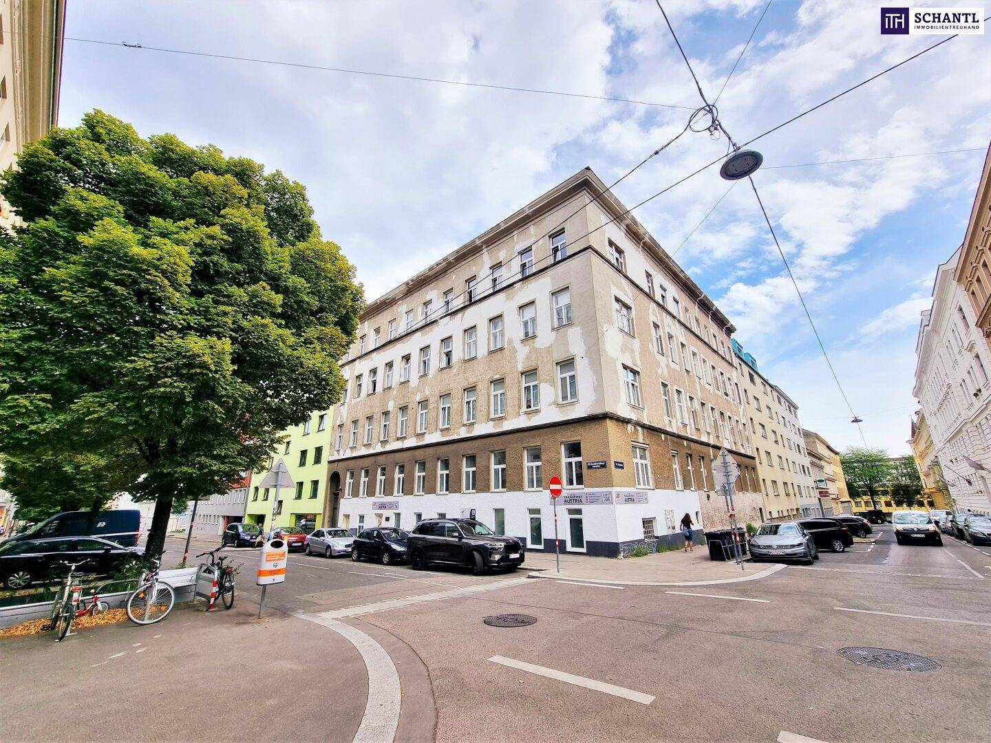 Eckgebäude an einer städtischen Kreuzung mit parkenden Autos und Bäumen, unter blauem Himmel.