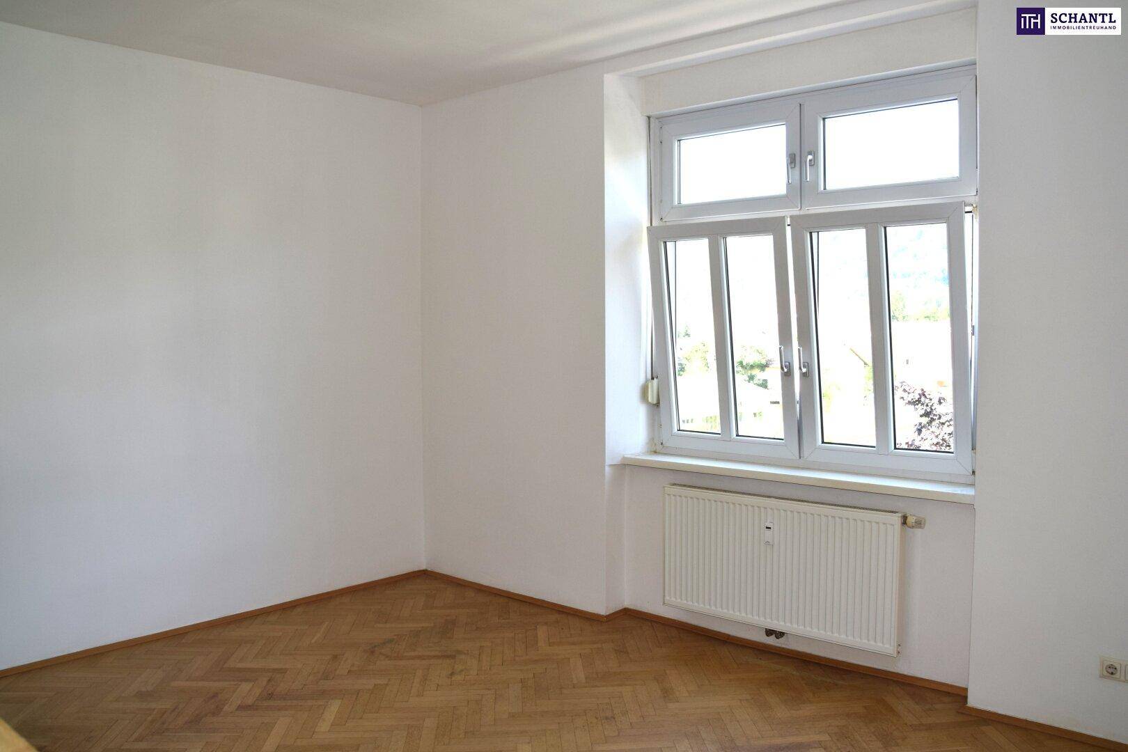 Helles Zimmer mit Fischgrätparkett, weißen Wänden und einem großen Fenster, das viel Licht hereinlässt.