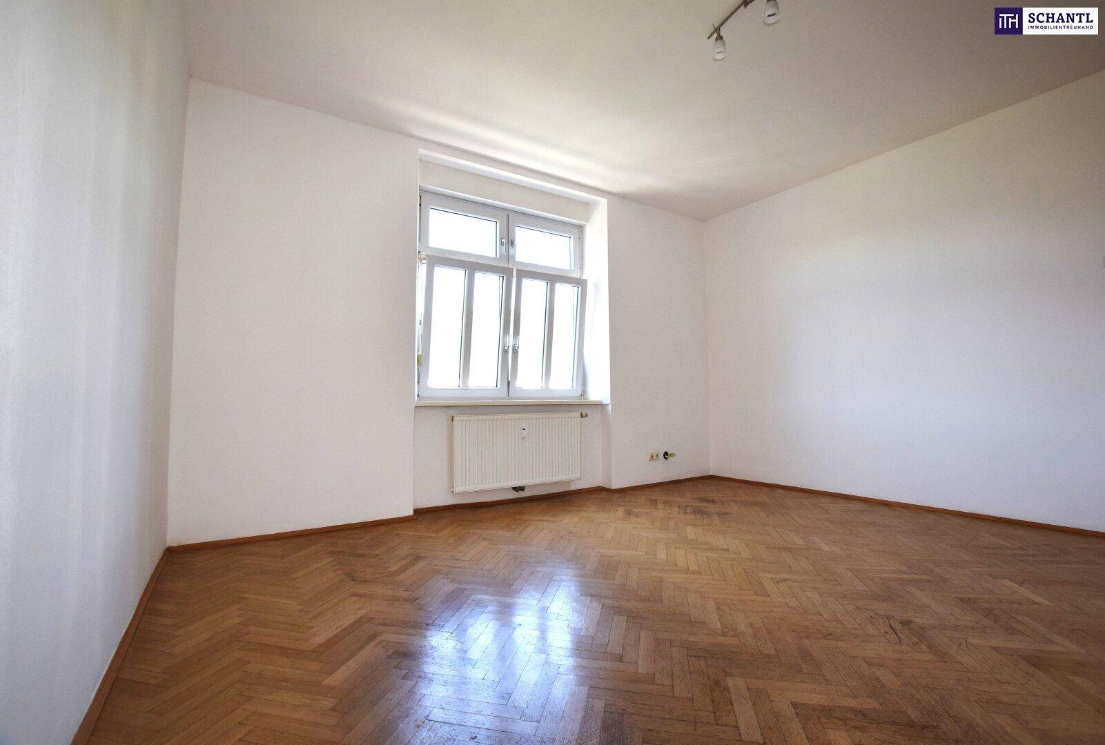 Helles, leeres Zimmer mit Fischgrätparkettboden, weißen Wänden und einem großen Fenster mit Heizkörper.