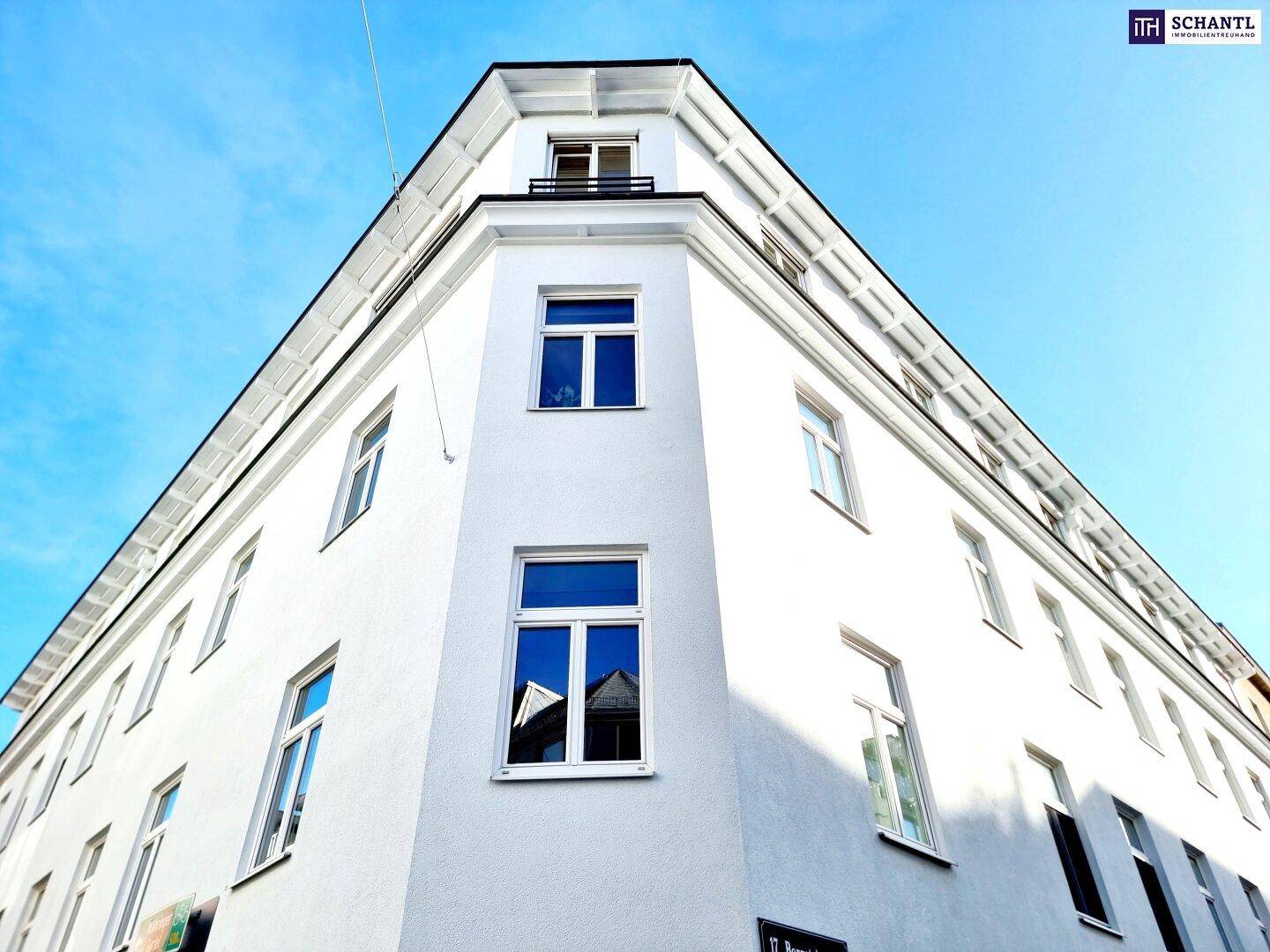 Weiße Eckfassade des Gebäudes mit blauen Fenstern unter klarem Himmel.