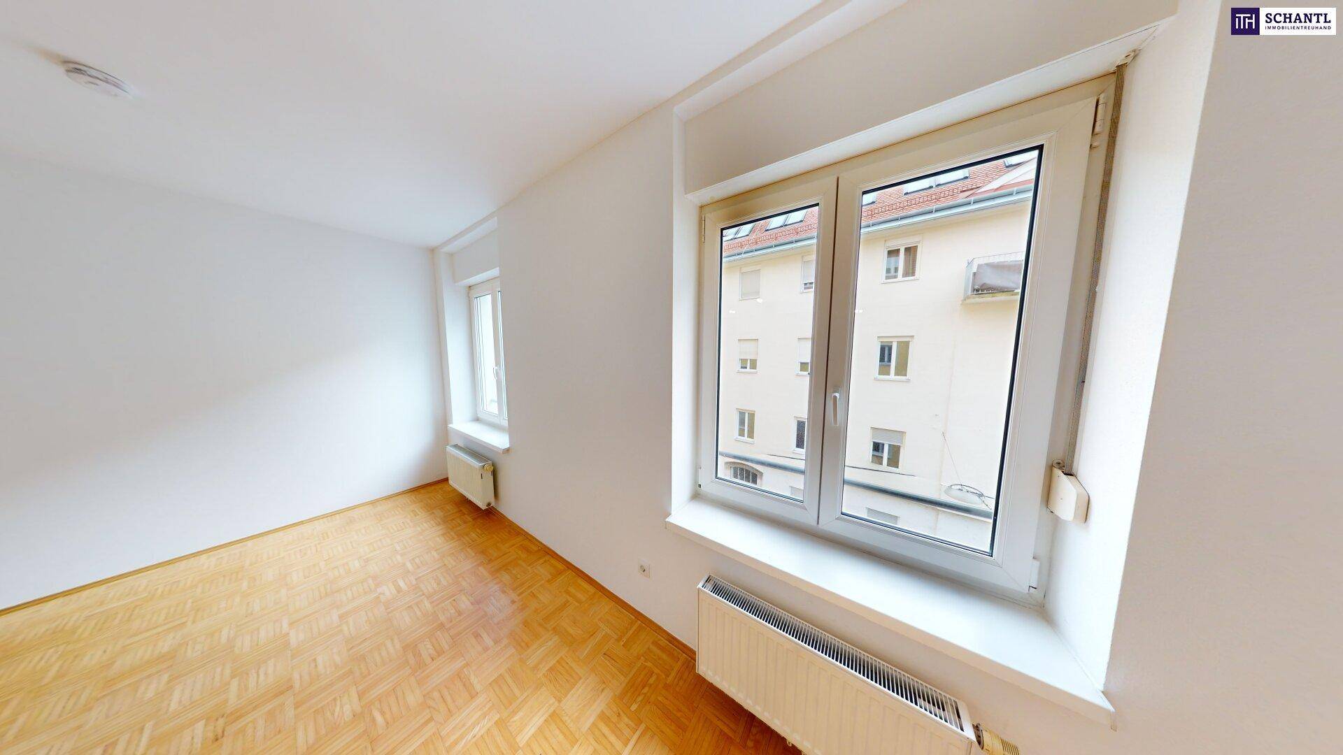Helles Zimmer mit Parkettboden und zwei Fenstern, die einen Blick auf die umliegenden Gebäude bieten.