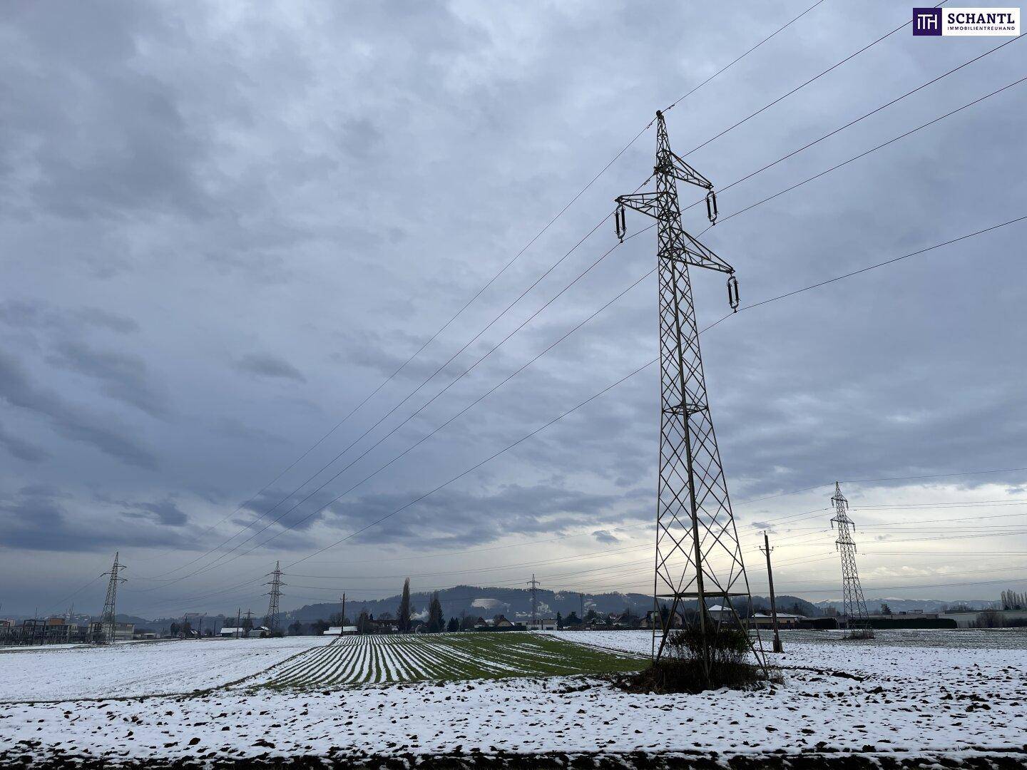 Blick über schneebedeckte Felder mit einem großen Strommast im Vordergrund unter einem dramatischen Wolkenhimmel.