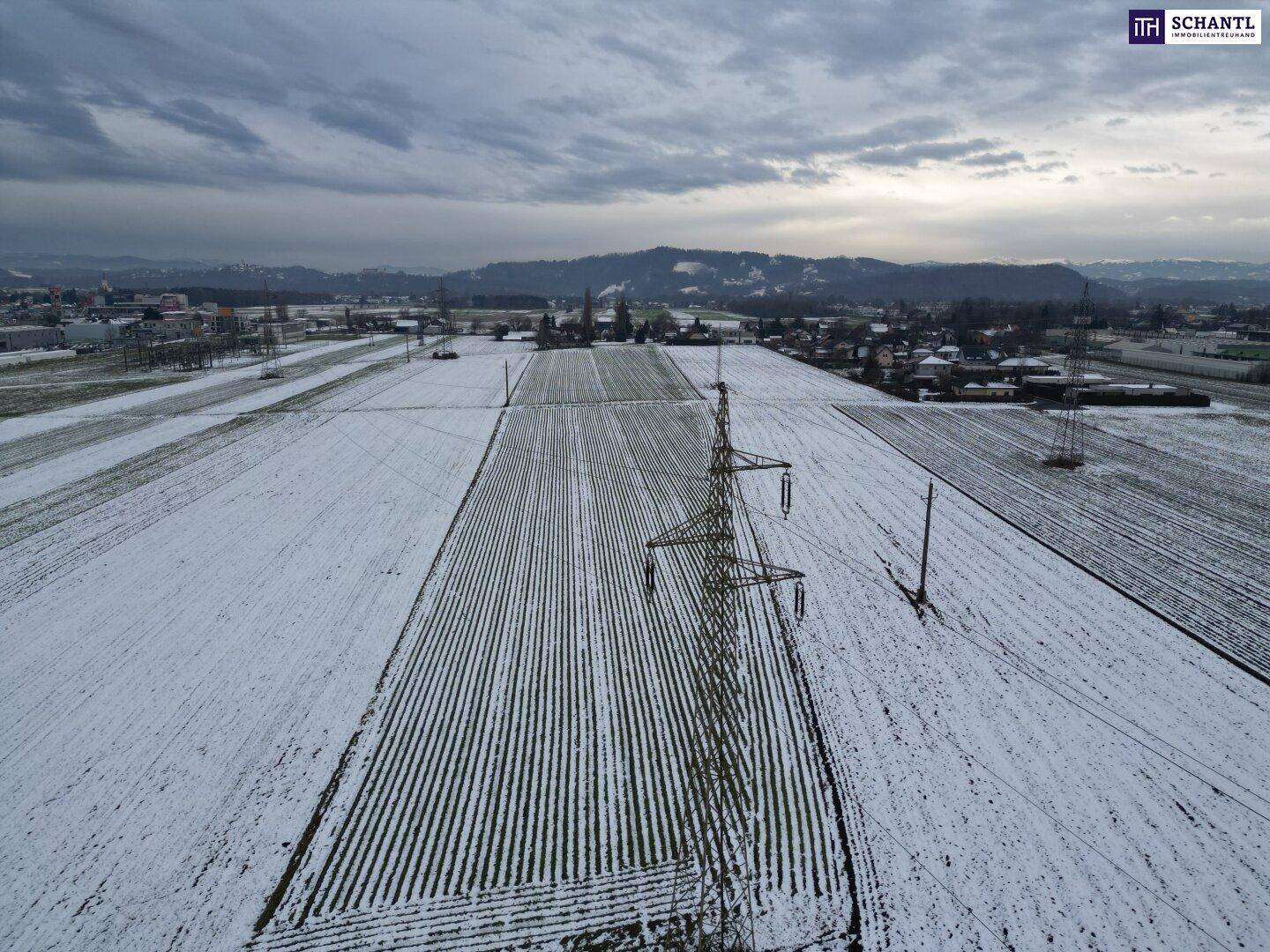 Panoramische Luftansicht von verschneiten Feldern mit Strommasten und einer Siedlung am Horizont unter grauem Himmel.