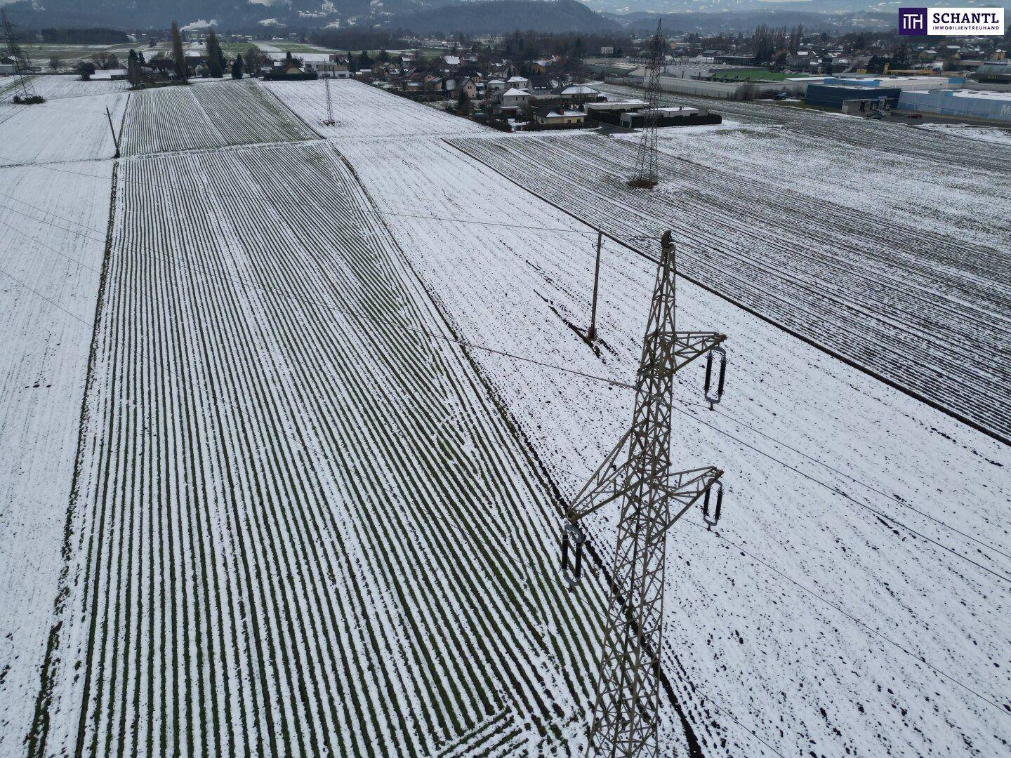 Luftaufnahme von schneebedeckten Feldern mit Strommasten und ländlicher Umgebung unter bewölktem Himmel.