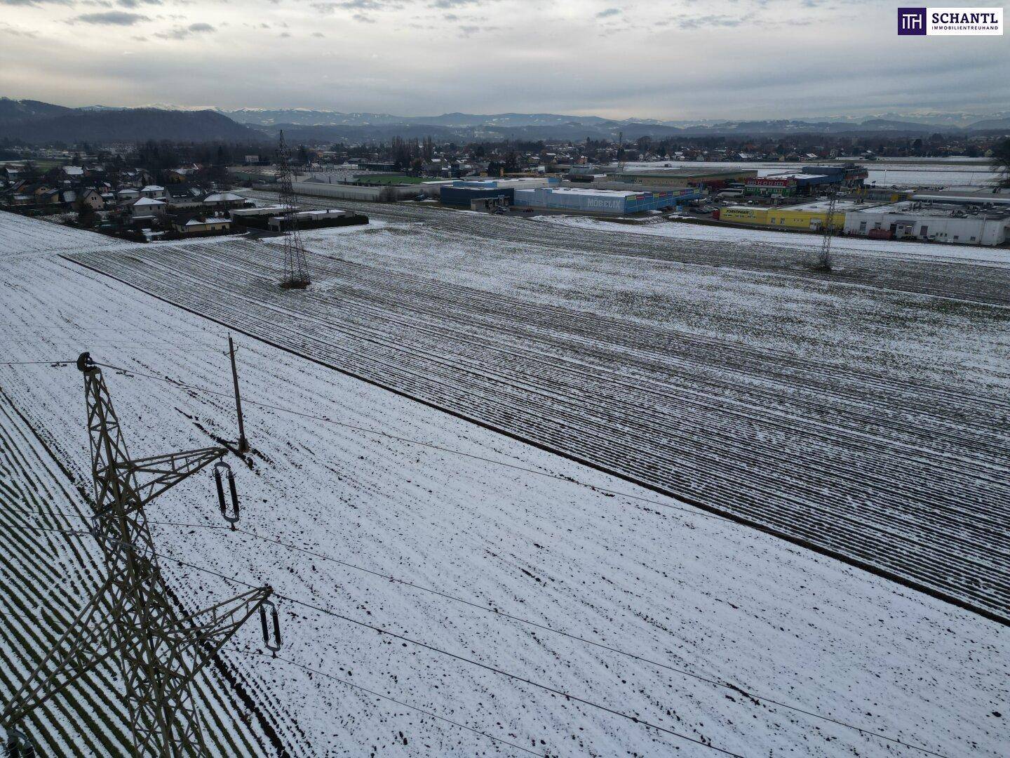 Weitläufige Luftaufnahme von schneebedeckten Ackerflächen mit Stromleitungen und Industriegebäuden in der Ferne.