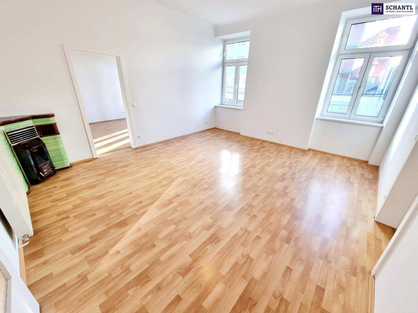 Helles Wohnzimmer mit Laminatboden, hohen Fenstern und einem traditionellen Ofen.