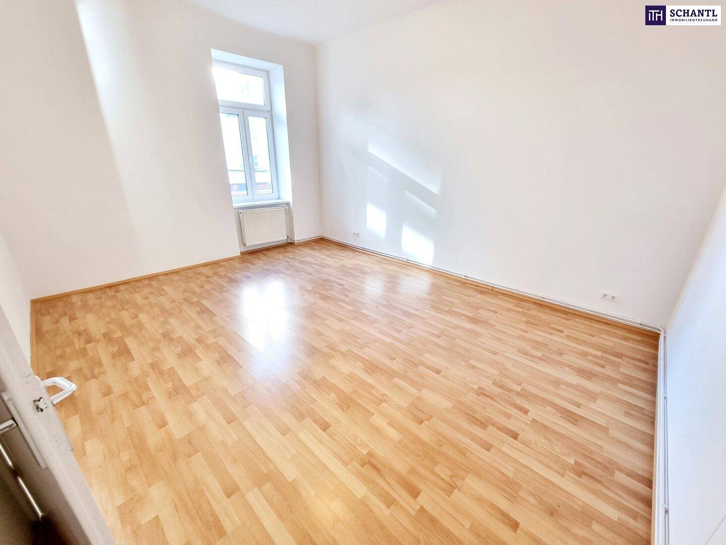 Leeres Zimmer mit Laminatboden, weißen Wänden und einem Fenster, das Tageslicht spendet.