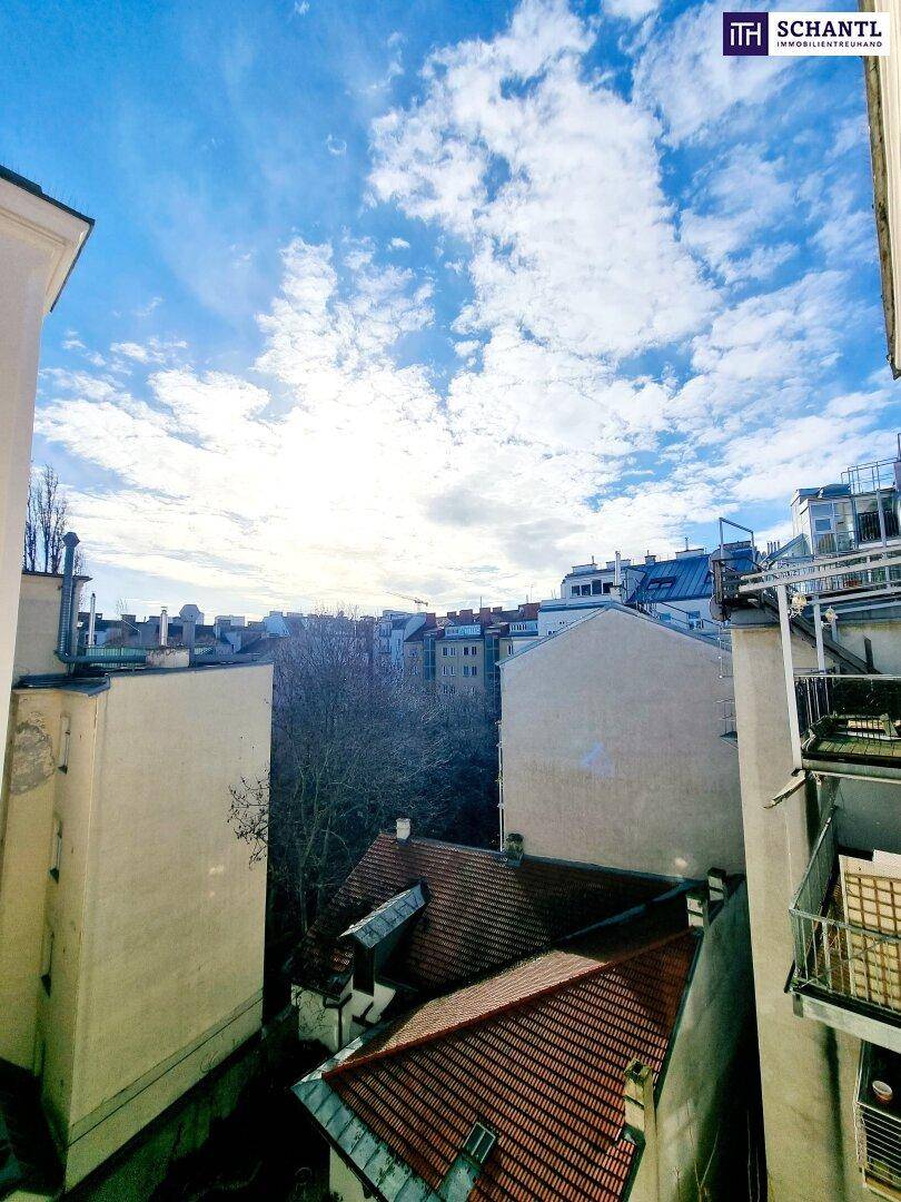 Blick aus einem oberen Stockwerk auf die Dächer benachbarter Gebäude, einen Innenhof und die Stadtkulisse unter blauem Himmel.