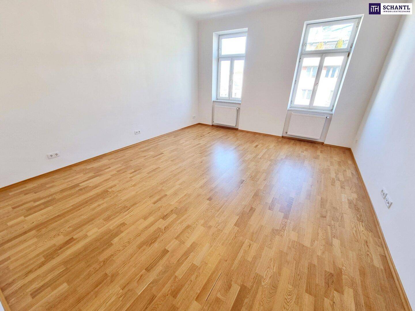 Geräumiger Raum mit hellem Parkettboden und zwei Fenstern, die viel Tageslicht hereinlassen.