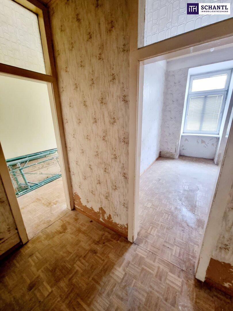 Blick durch eine Tür in ein weiteres Zimmer mit altem Parkettboden und abgenutzter Tapete.