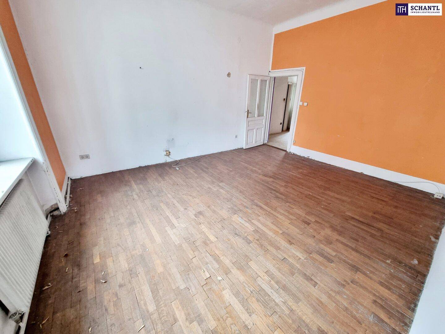 Helles Zimmer mit Fischgrätparkett, großem Fenster und einer orangefarbenen Wand.
