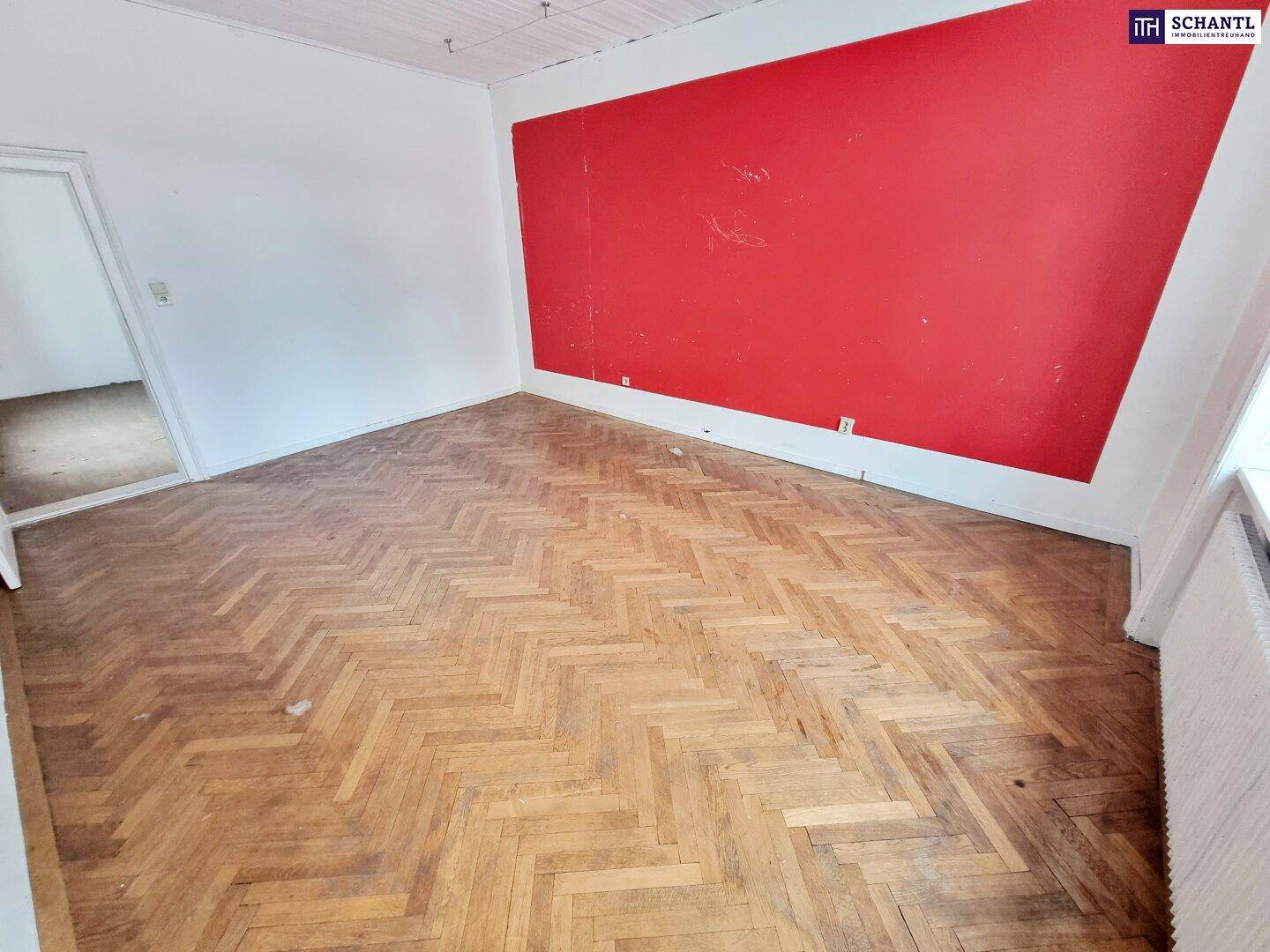 Geräumiges Zimmer mit Fischgrätparkett, großem Fenster und einer markanten roten Wand.