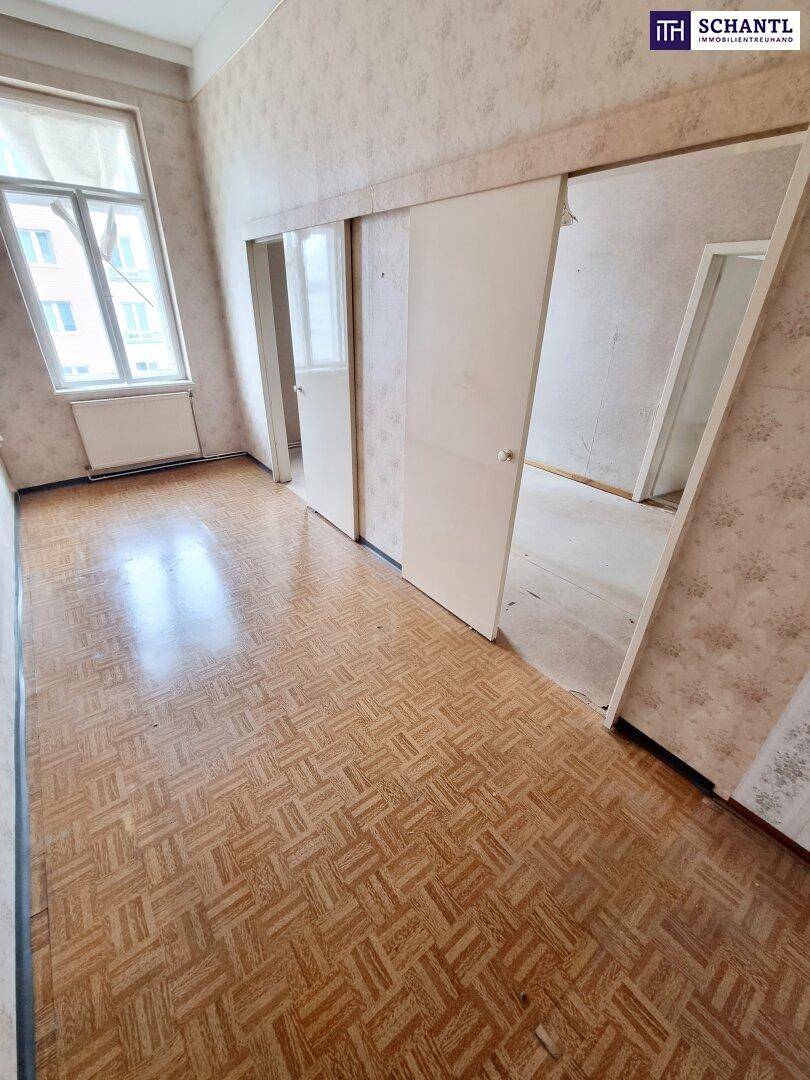 Renovierungsbedürftiger Raum mit gemusterter Tapete, altem Parkettboden und großem Fenster.