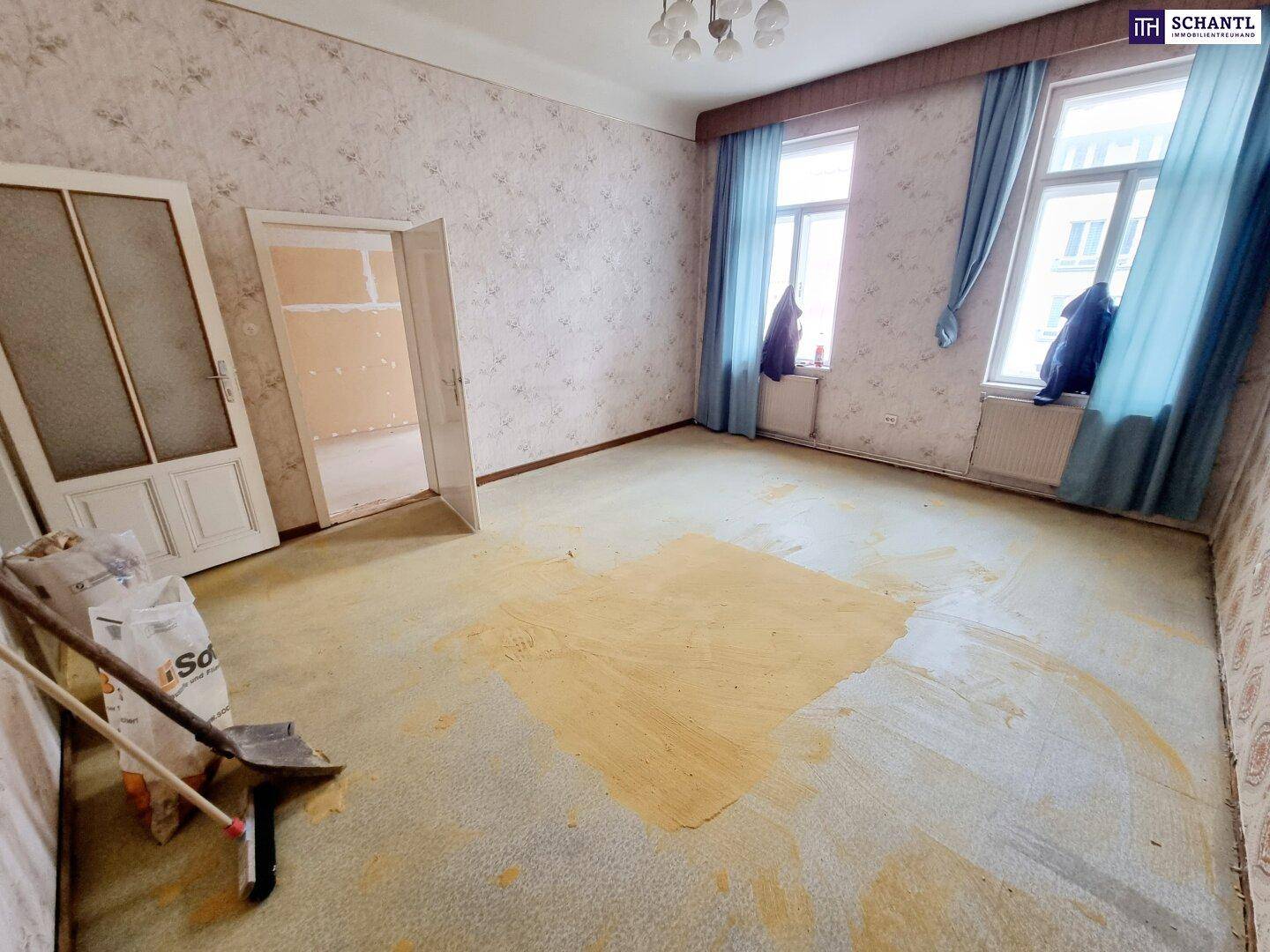 Renovierungsbedürftiger Raum mit alten Tapeten, Teppichboden und Fenstern mit blauen Vorhängen.
