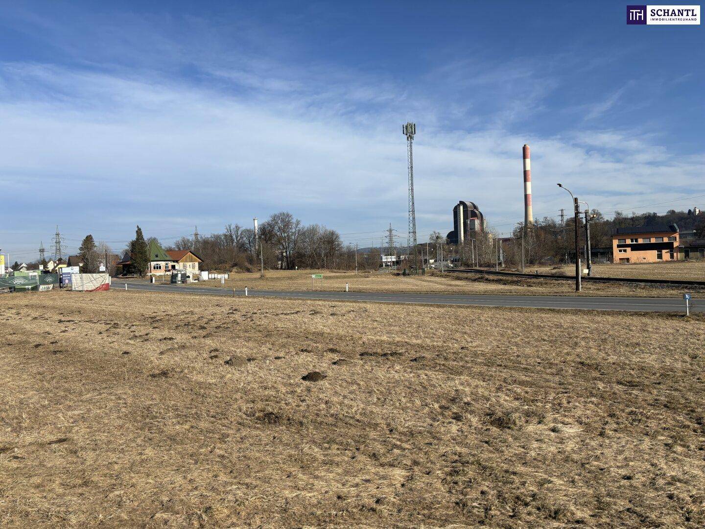 Weitläufiges trockenes Feld neben einer Straße mit industriellen Schornsteinen und Gebäuden in der Ferne.